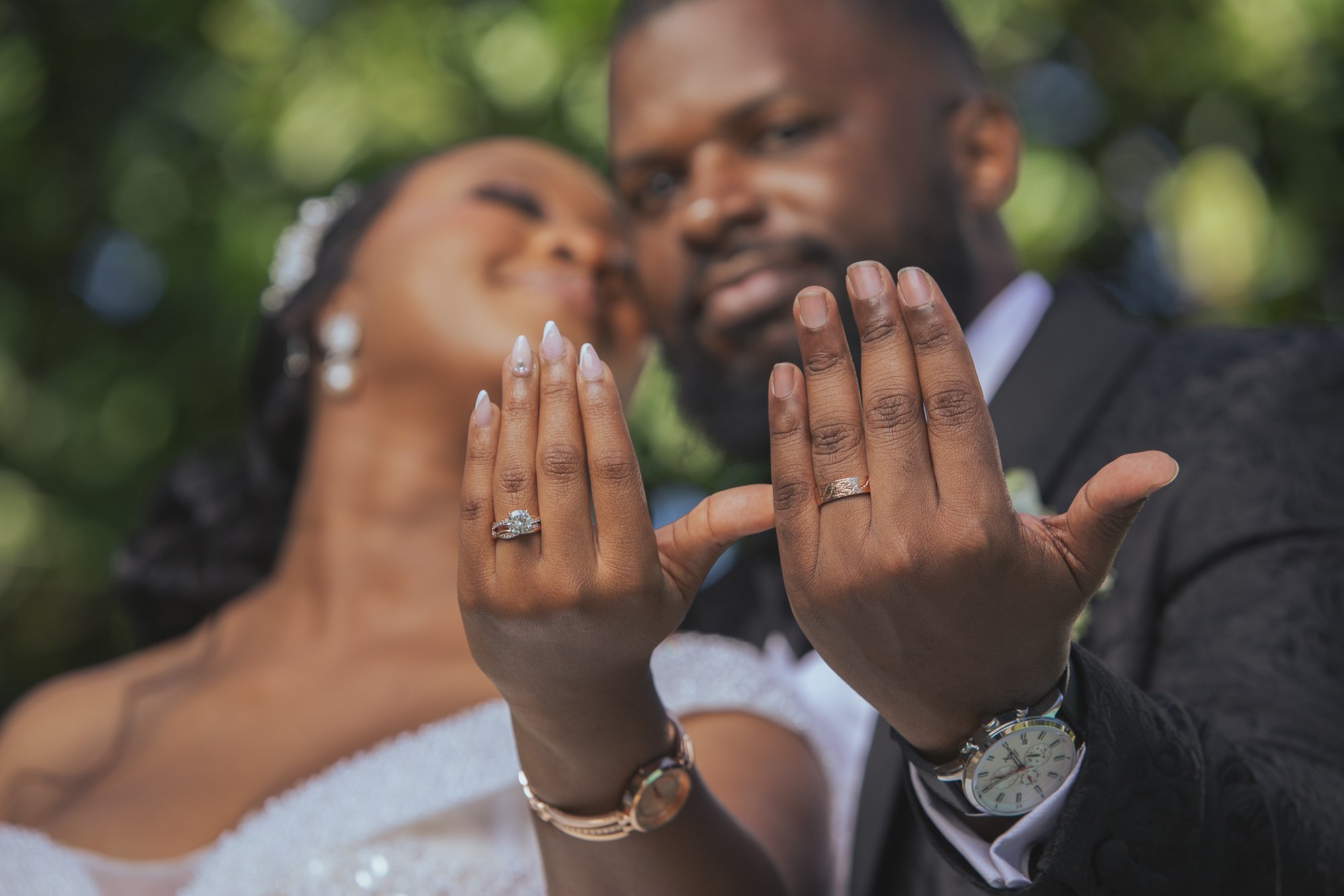 Close-up of a bride and groom showing their wedding rings on overlapping hands, with the bride smiling and the groom looking at her, outdoors with blurred green foliage in the background.