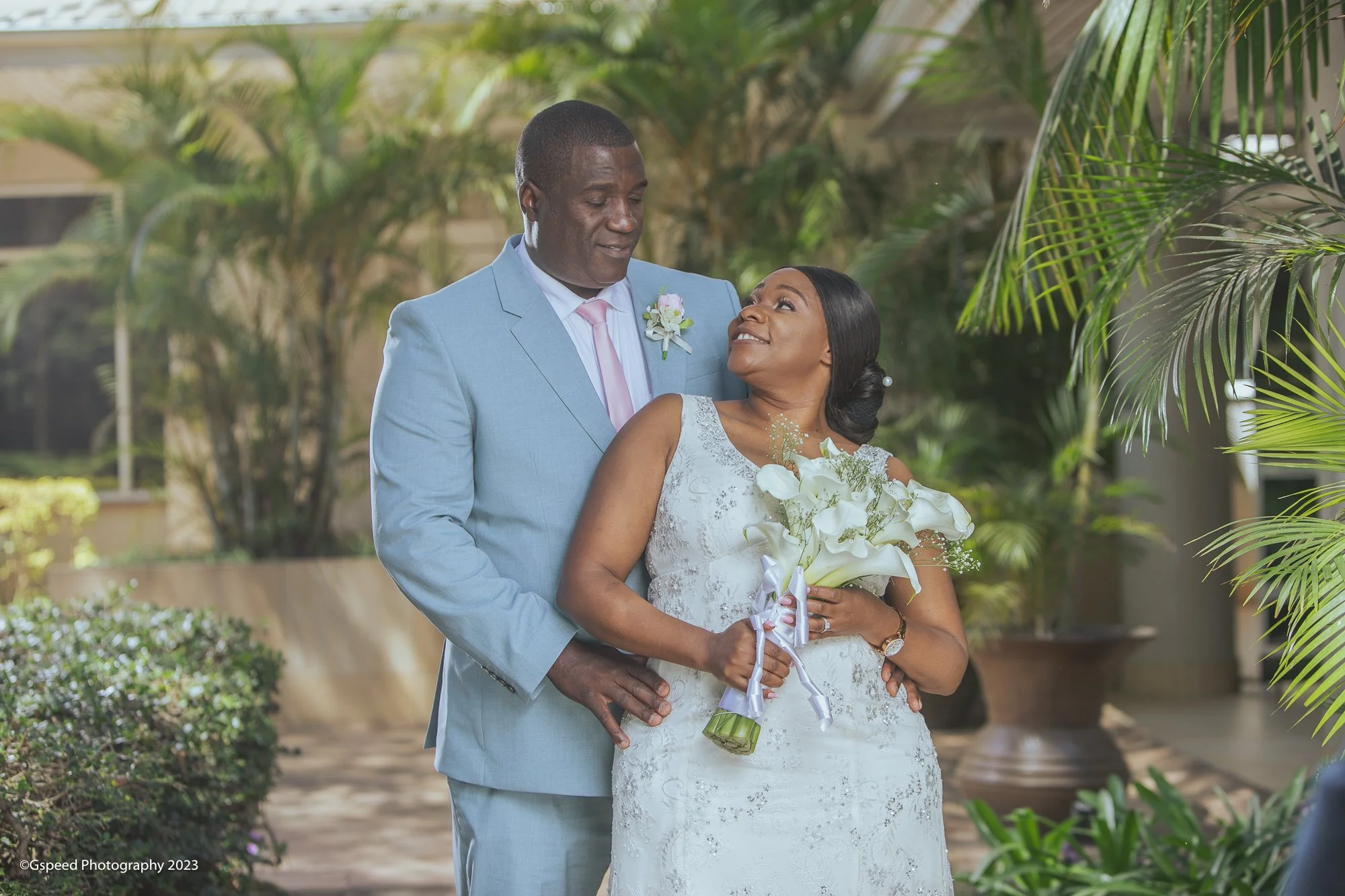 A couple in wedding attire focusing lovingly on each other outdoors surrounded by greenery, with the woman holding a bouquet of white flowers.