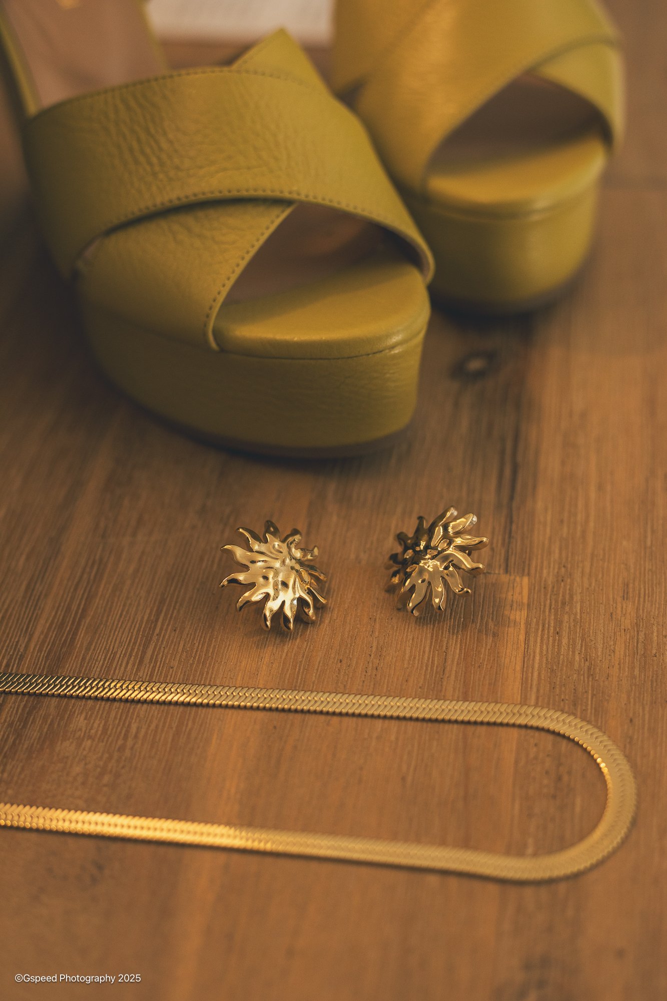 Close-up of a pair of gold earrings with a leaf design, placed on a wooden surface with a gold zipper. A pair of yellow high-heeled shoes with a textured surface are in the background.
