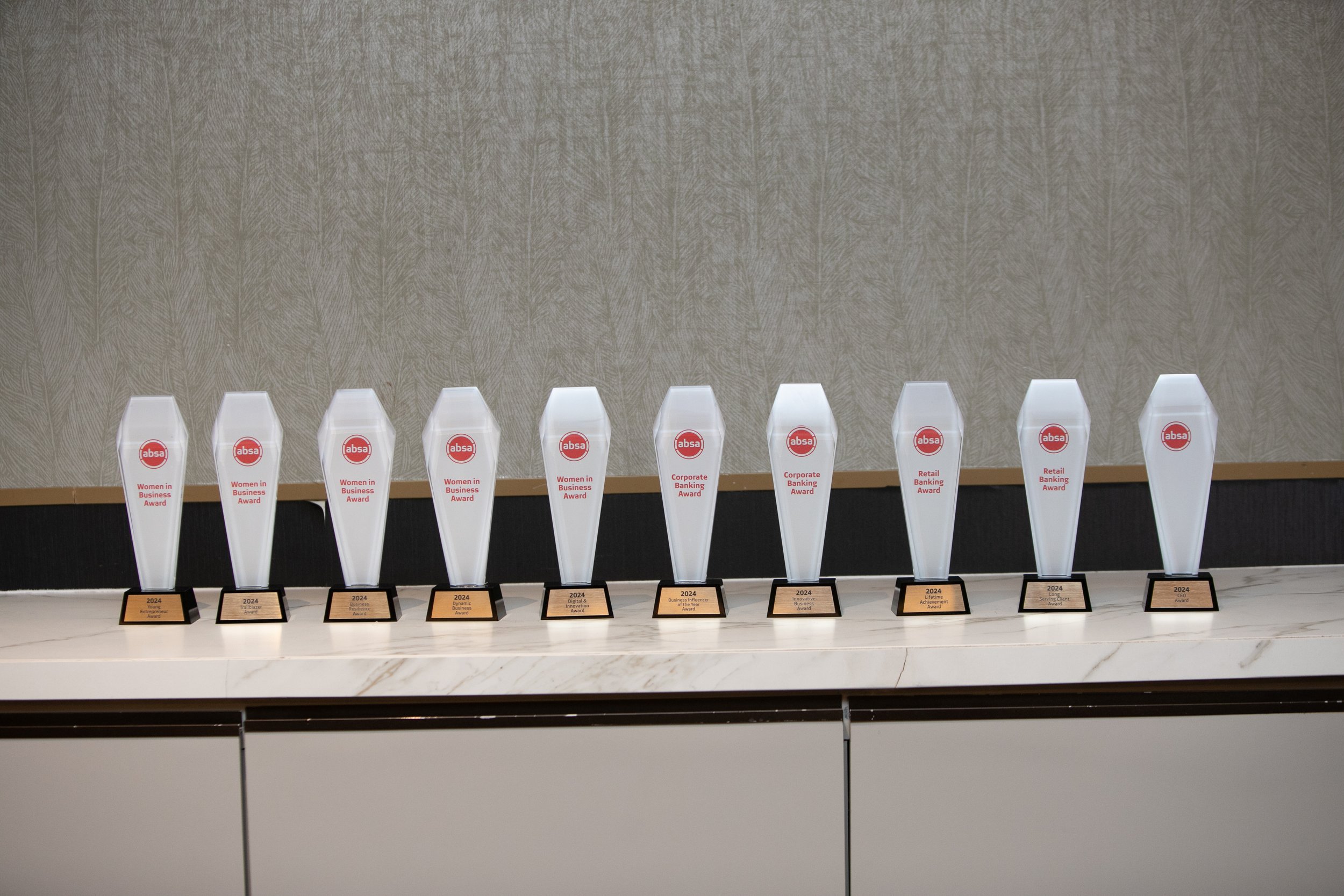 A row of ten clear awards with red logos and text, each with a black base, displayed on a white marble counter against a textured beige wall