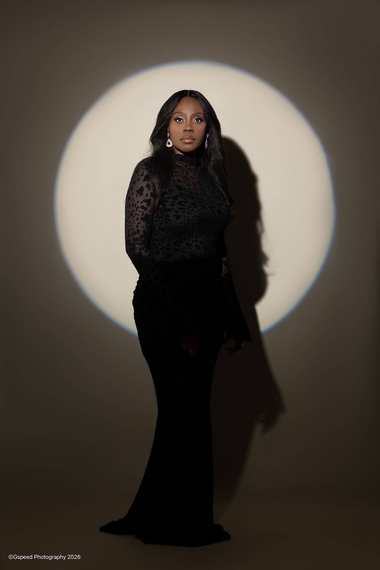 A woman standing in front of a circular spotlight on a dark background, wearing a black patterned top with long sleeves, black pants, and large earrings.