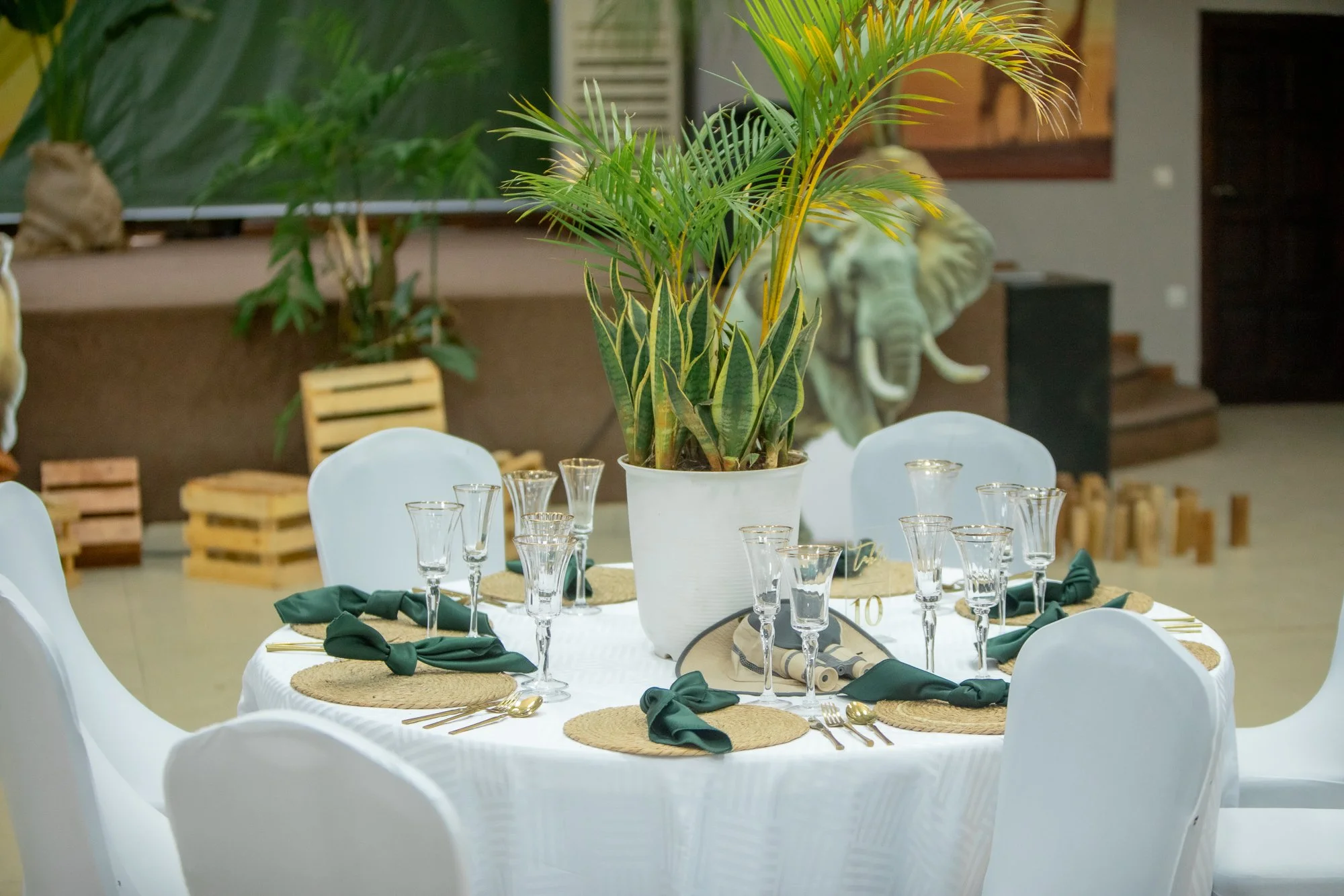 Elegant round table with white tablecloth, green napkins, glassware, and a central potted plant, set for a formal event in a decorated venue.