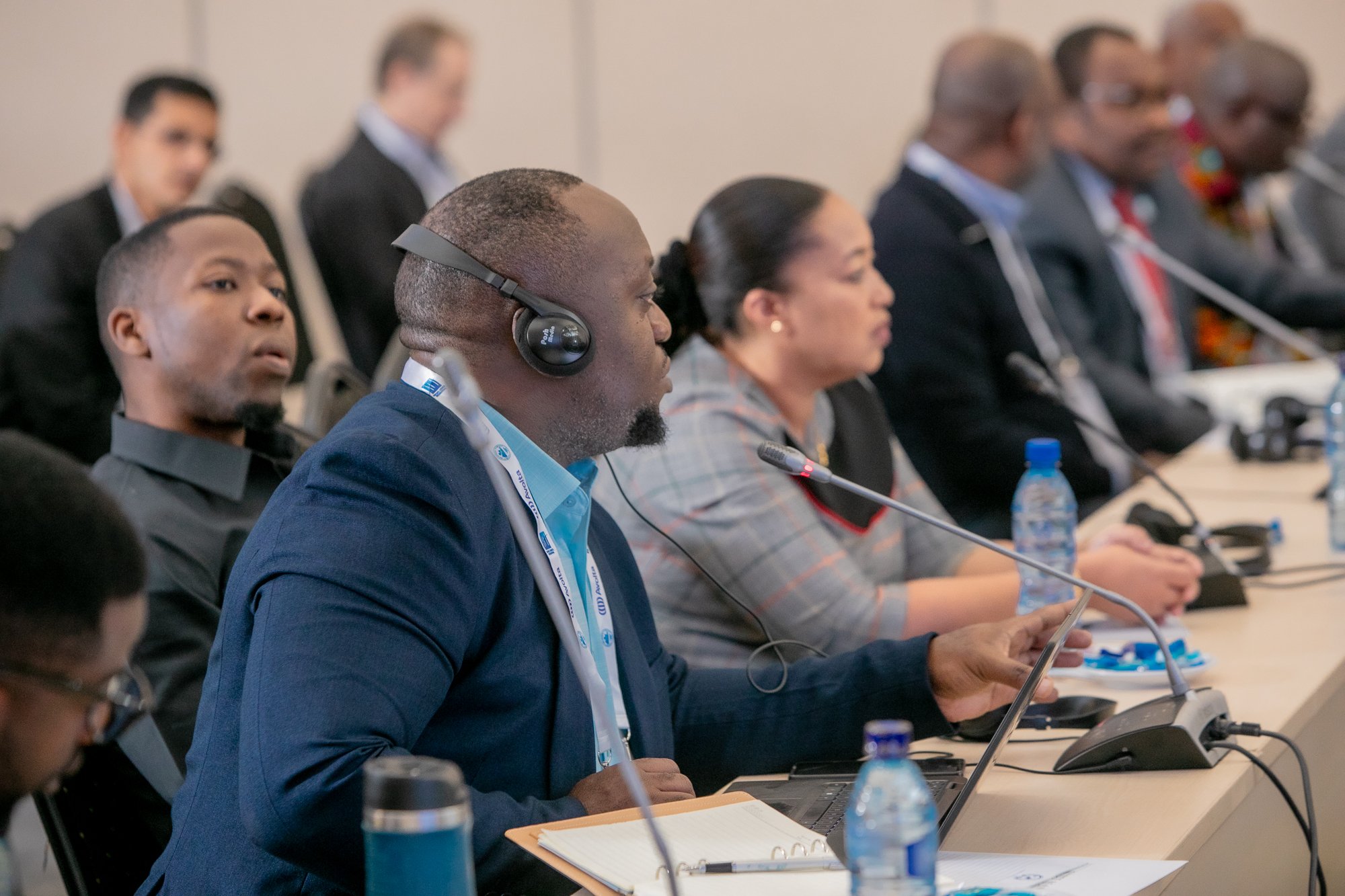 A group of people sitting in rows at a conference or meeting, listening attentively, some with headsets, taking notes, and looking towards the front of the room.
