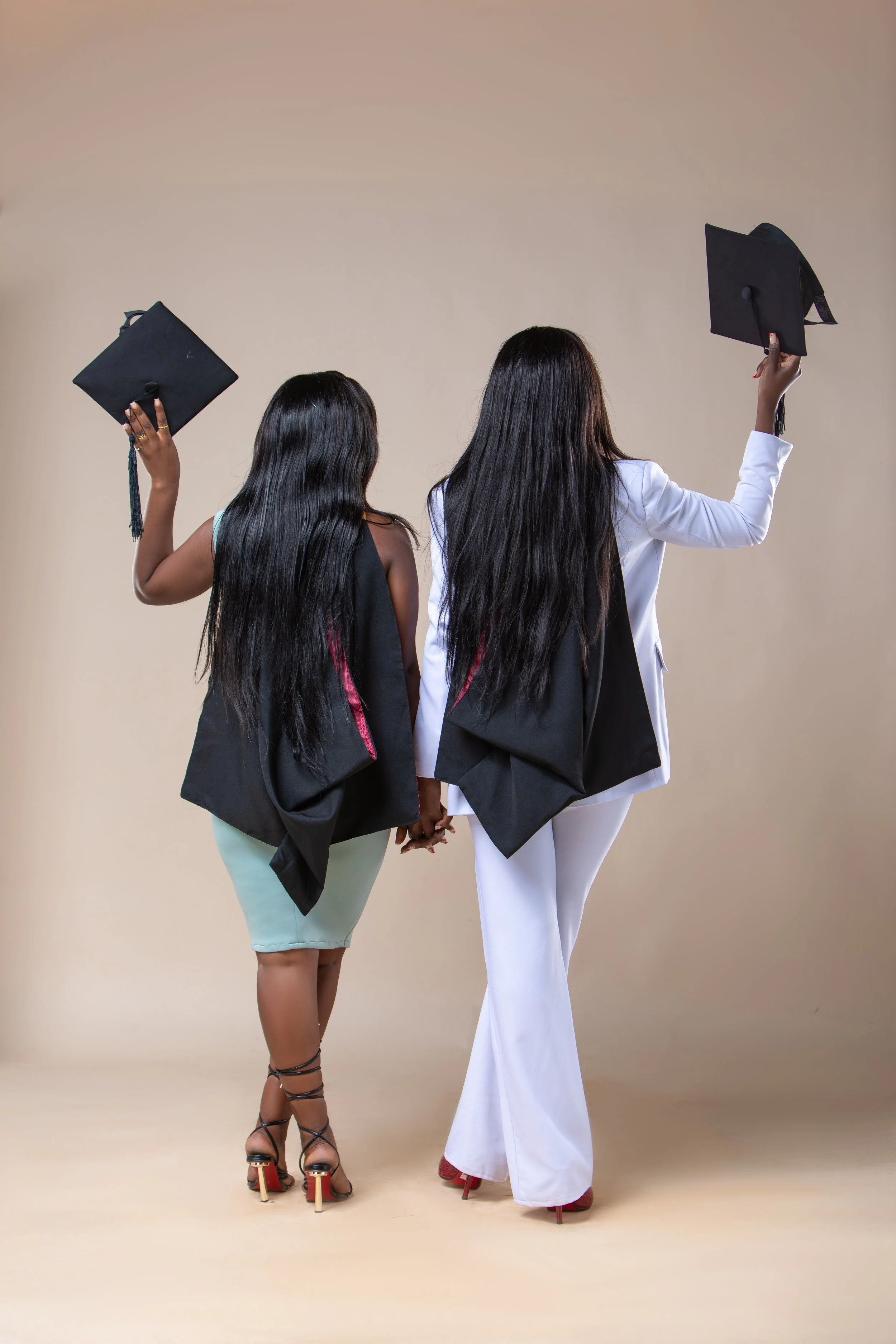 Two women dressed in graduation gowns, holding hands, with their backs facing the camera. One woman is wearing a black gown with a light blue dress underneath and strappy high heels, while the other woman is wearing a white gown with red shoes.