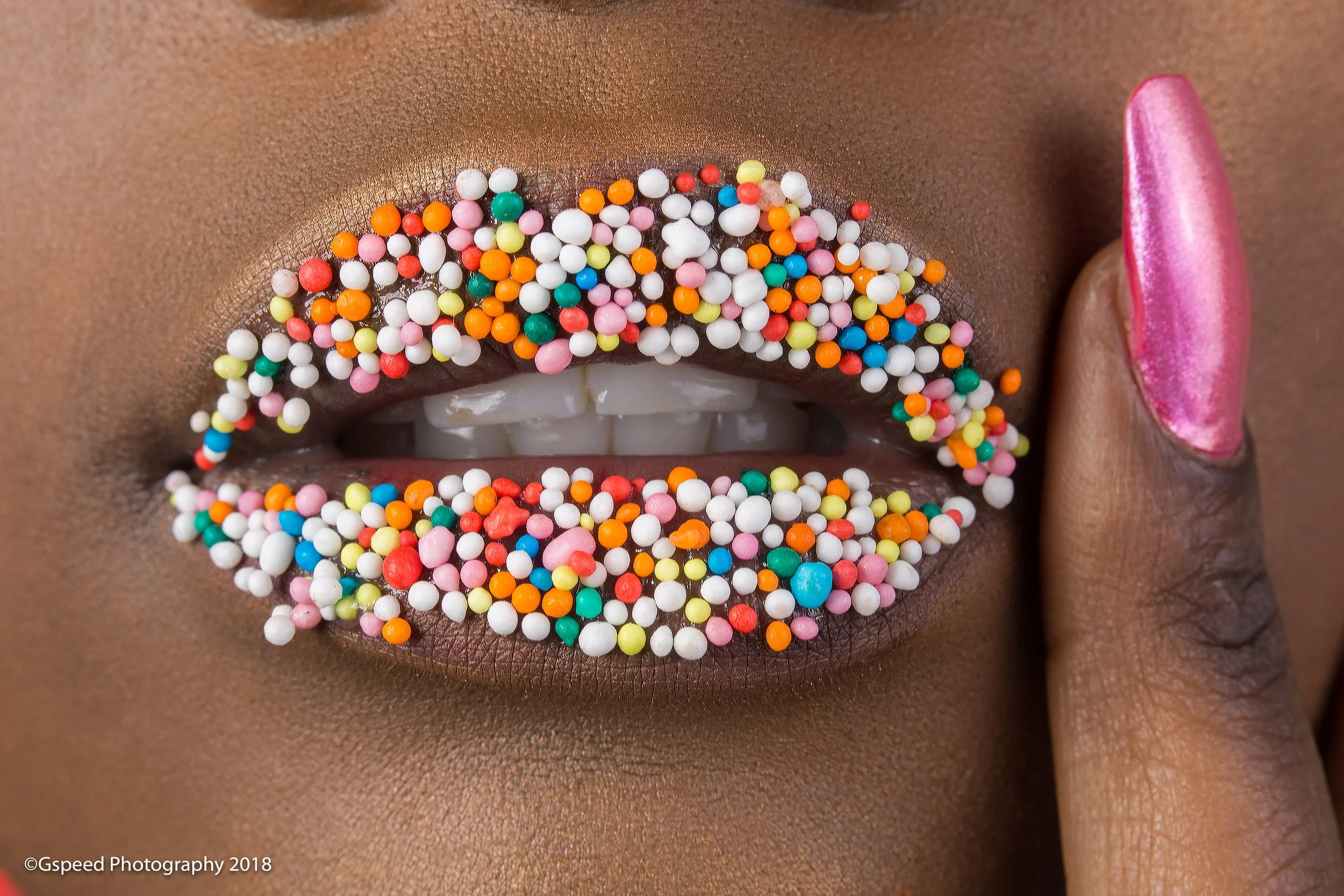 Close-up of a person's lips with pink lipstick, decorated with colorful sprinkles and sugar beads, with a finger touching the lips.
