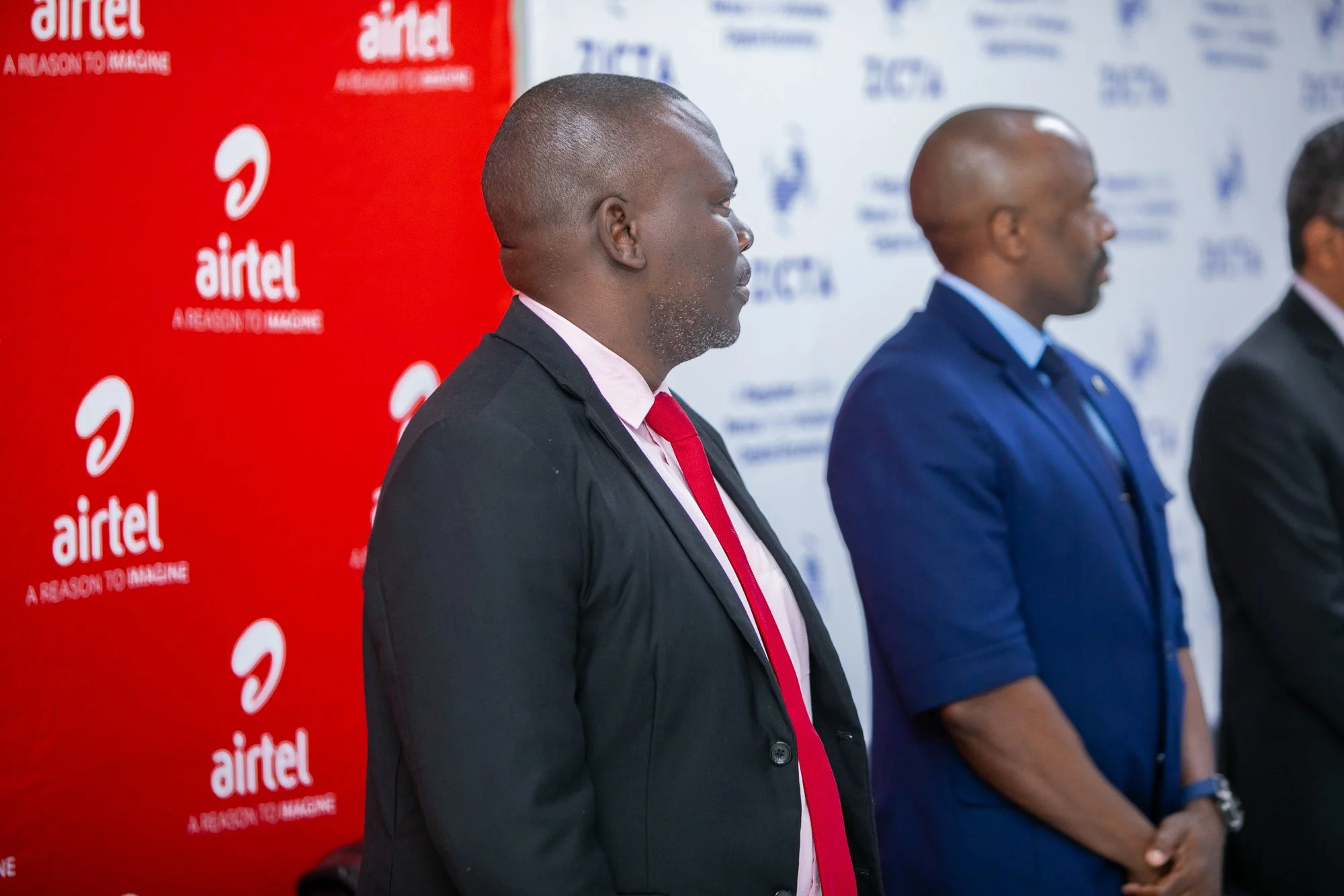 A man in a black suit and pink tie, with a shaved head and gray beard, standing in profile. Behind him, a man in a blue suit and a bald head, and part of another man in a black suit with glasses are visible. The background features a red Airtel banne