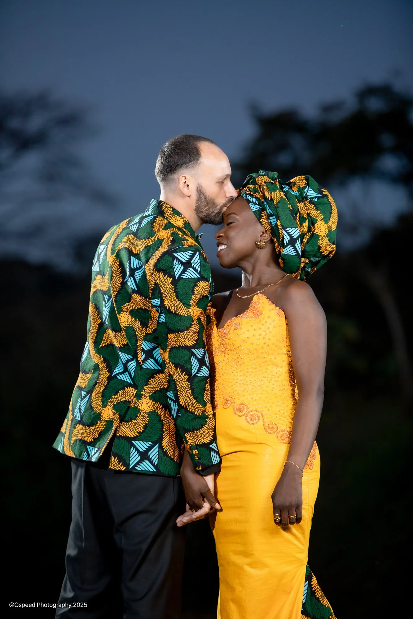 A couple standing close together, holding hands, with the man kissing the woman's forehead. Both are dressed in vibrant, traditional African attire, with the woman wearing a yellow dress and headwrap, and the man wearing a matching patterned suit jac