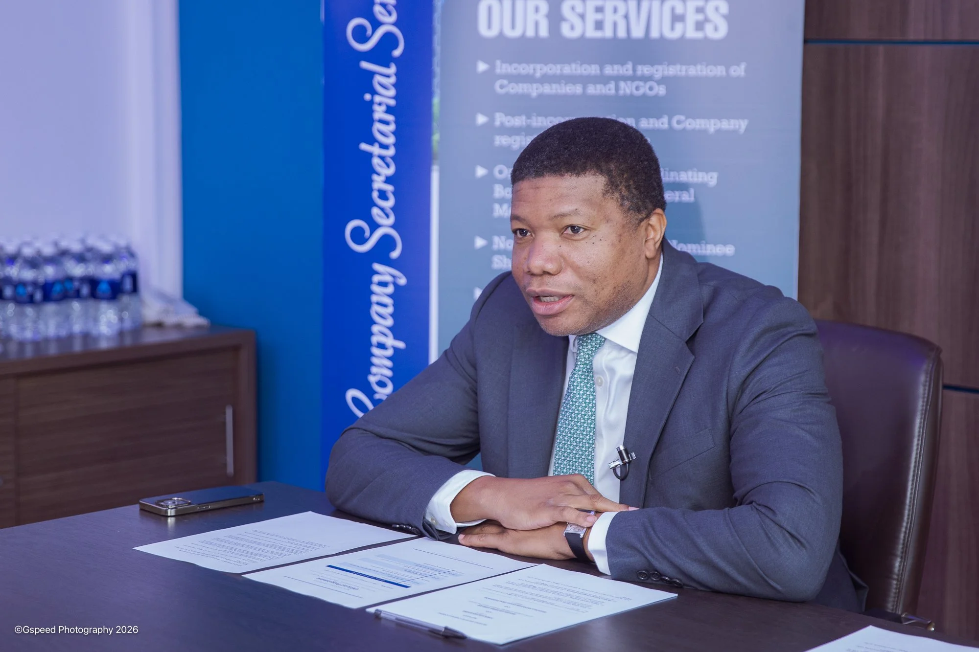A man in a suit sitting at a desk with documents, a pen, and a phone, in front of a blue and gray banner that reads 'Our Services' and 'Incorporation and registration of Companies and NGOs.'