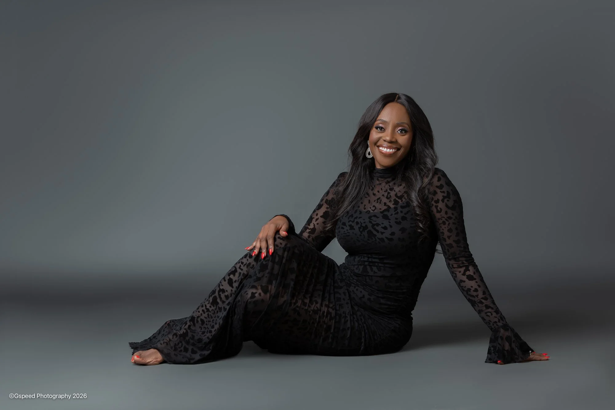 A smiling woman with long dark hair sitting on the floor against a gray background, wearing a black leopard print dress and gold earrings.