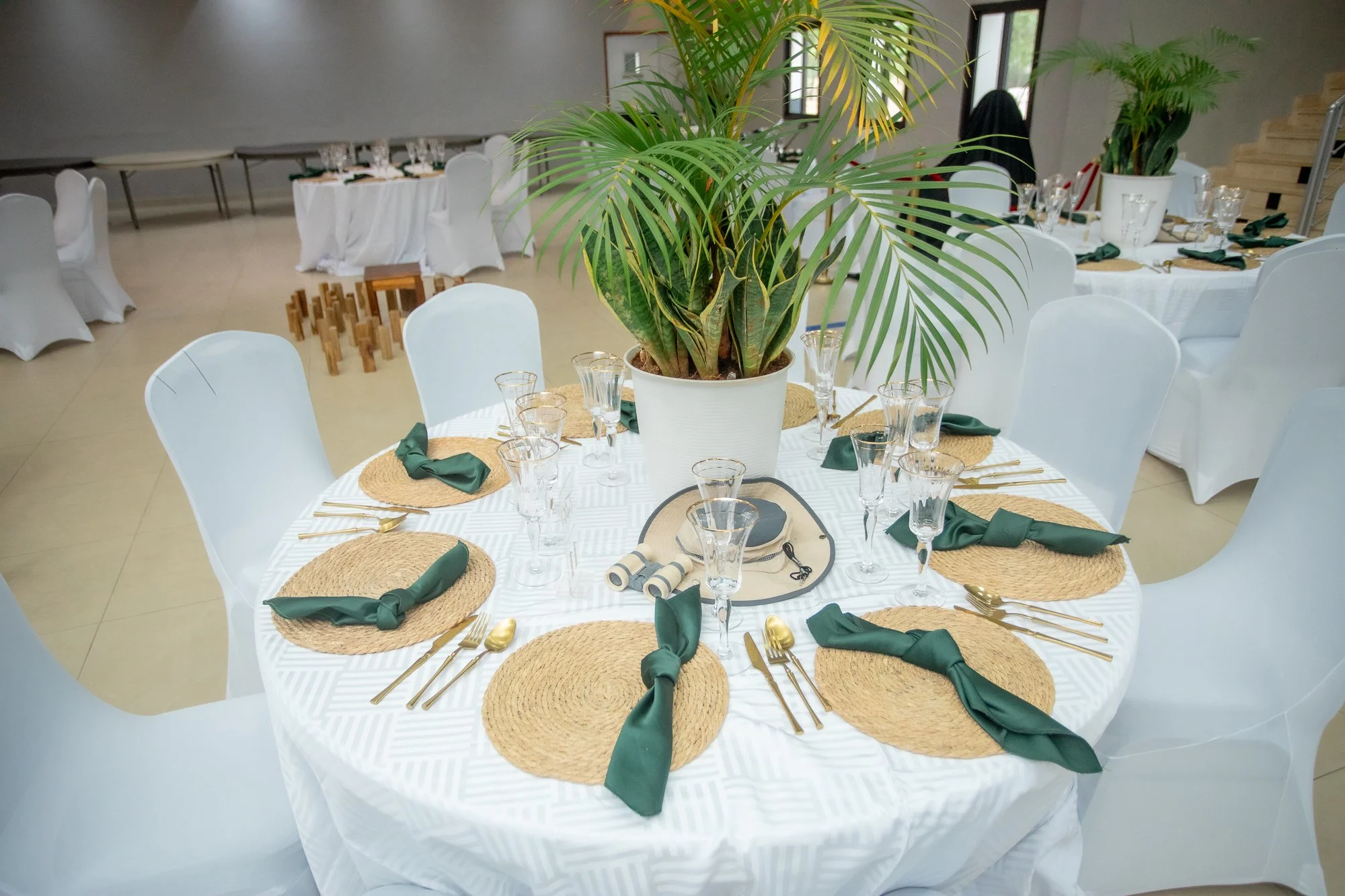 Round dining table decorated for an event, with white tablecloth, woven placemats, green cloth napkins, gold flatware, and glassware. A large potted palm plant is in the center of the table. White chairs surround the table, and additional set tables 