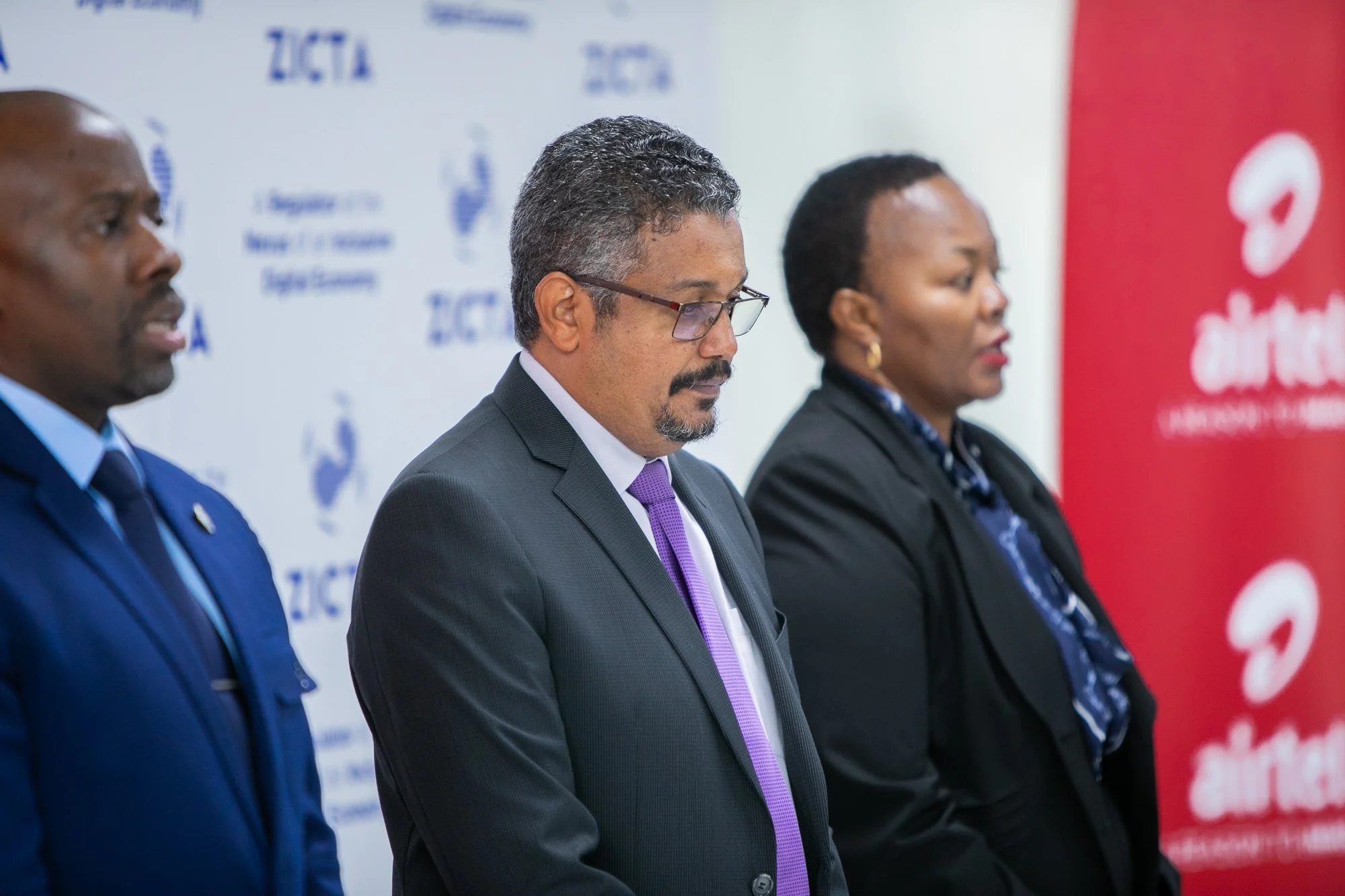Three people in formal attire standing side by side at a conference, with a background displaying logos and text.