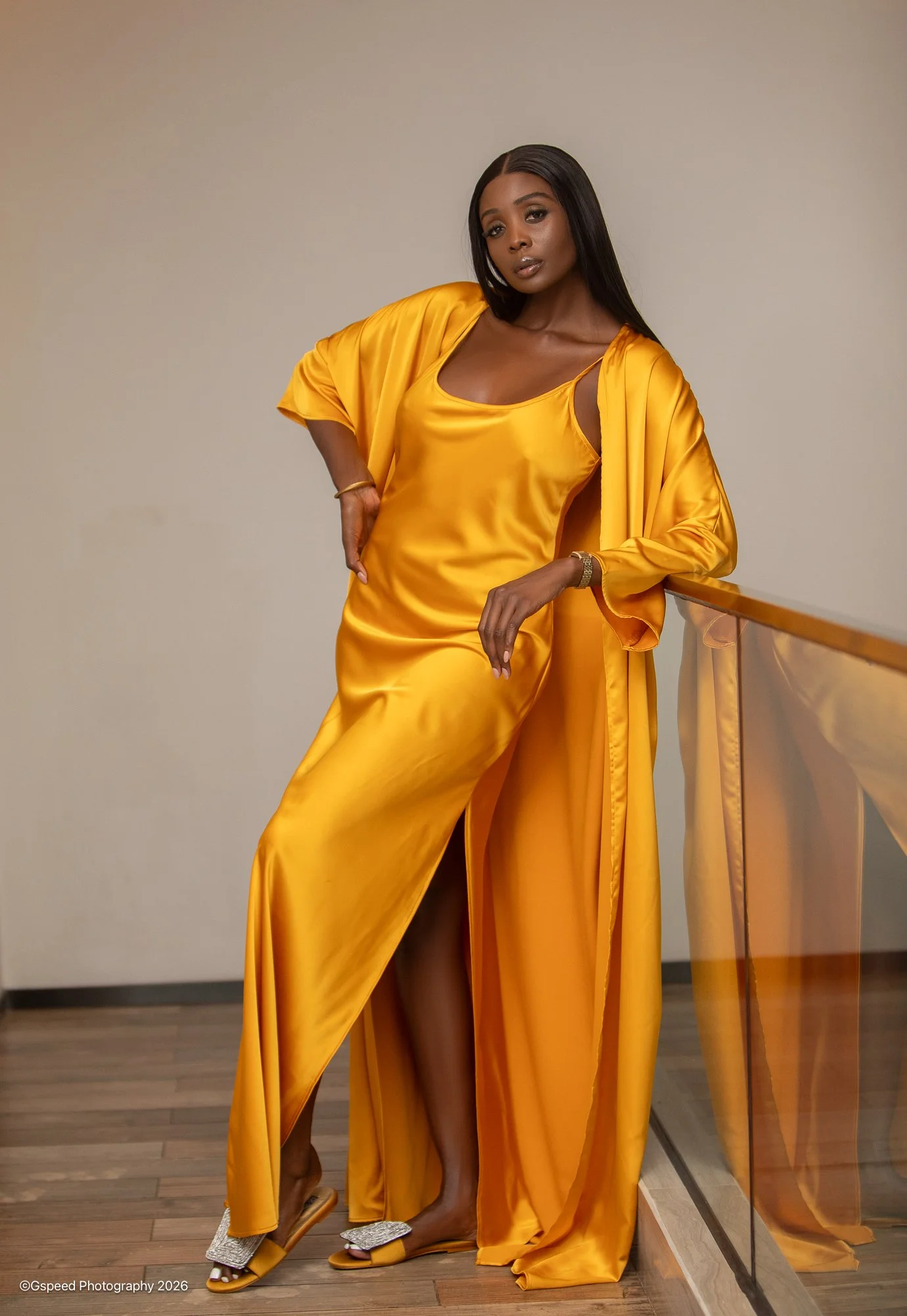 A woman in a flowing, loose-fitting satin yellow dress and matching long robe, standing indoors against a plain beige wall, with her right hand on her hip. She is wearing high-heeled sandals with rhinestone embellishments and gold accents, and has lo