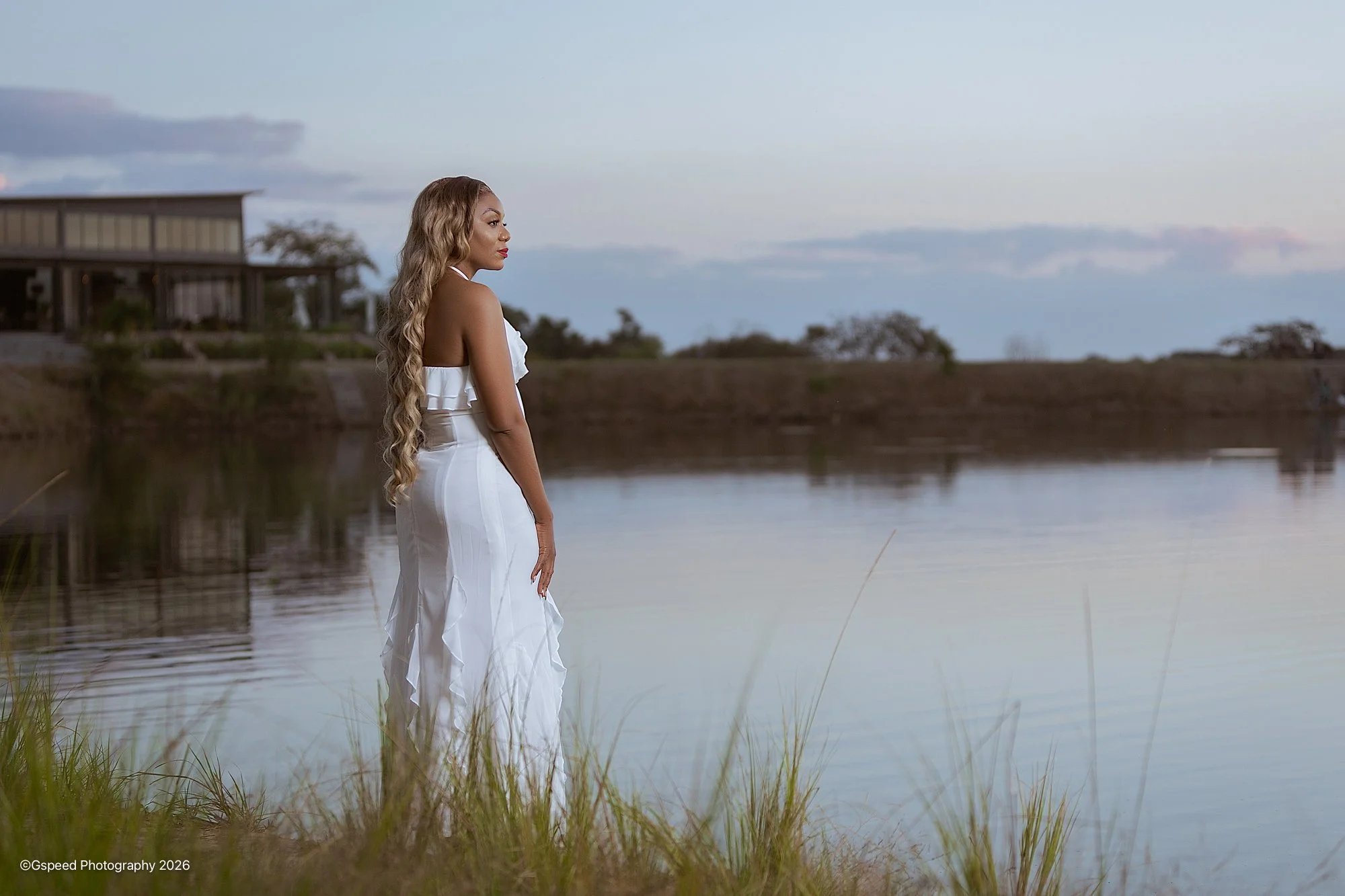 A woman with long, wavy blonde hair wearing a white dress stands by a calm body of water during dusk, with a modern house on stilts in the background and trees on the horizon.
