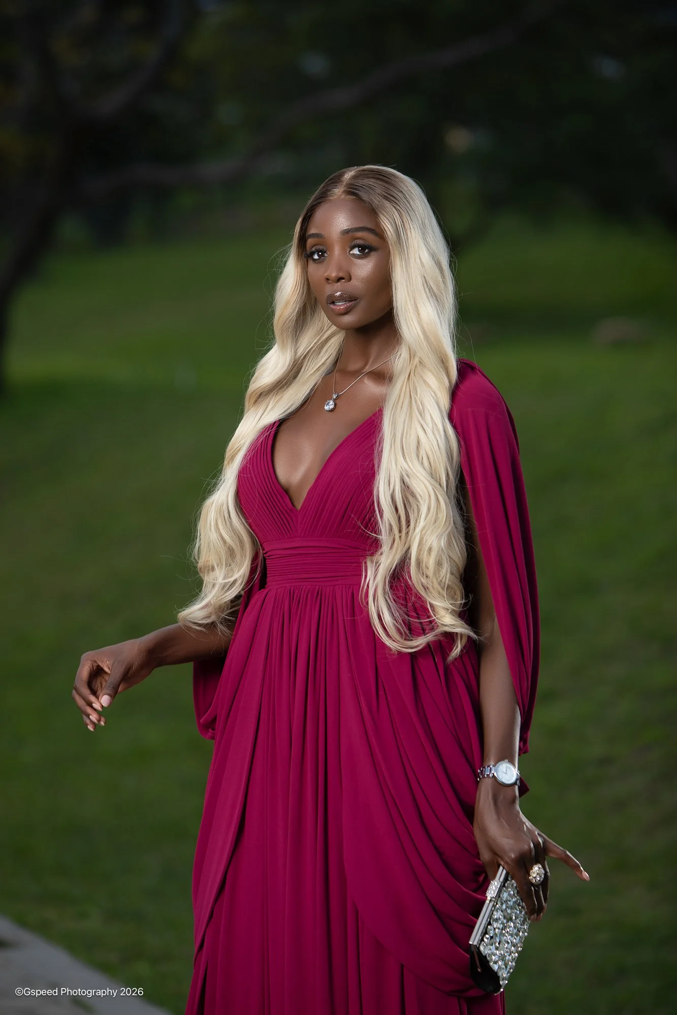 A woman in a deep red, pleated, floor-length gown stands outdoors in front of a blurred green background. She has long, styled platinum blonde hair and is wearing a necklace, watch, and rings. She is holding a bejeweled clutch purse.