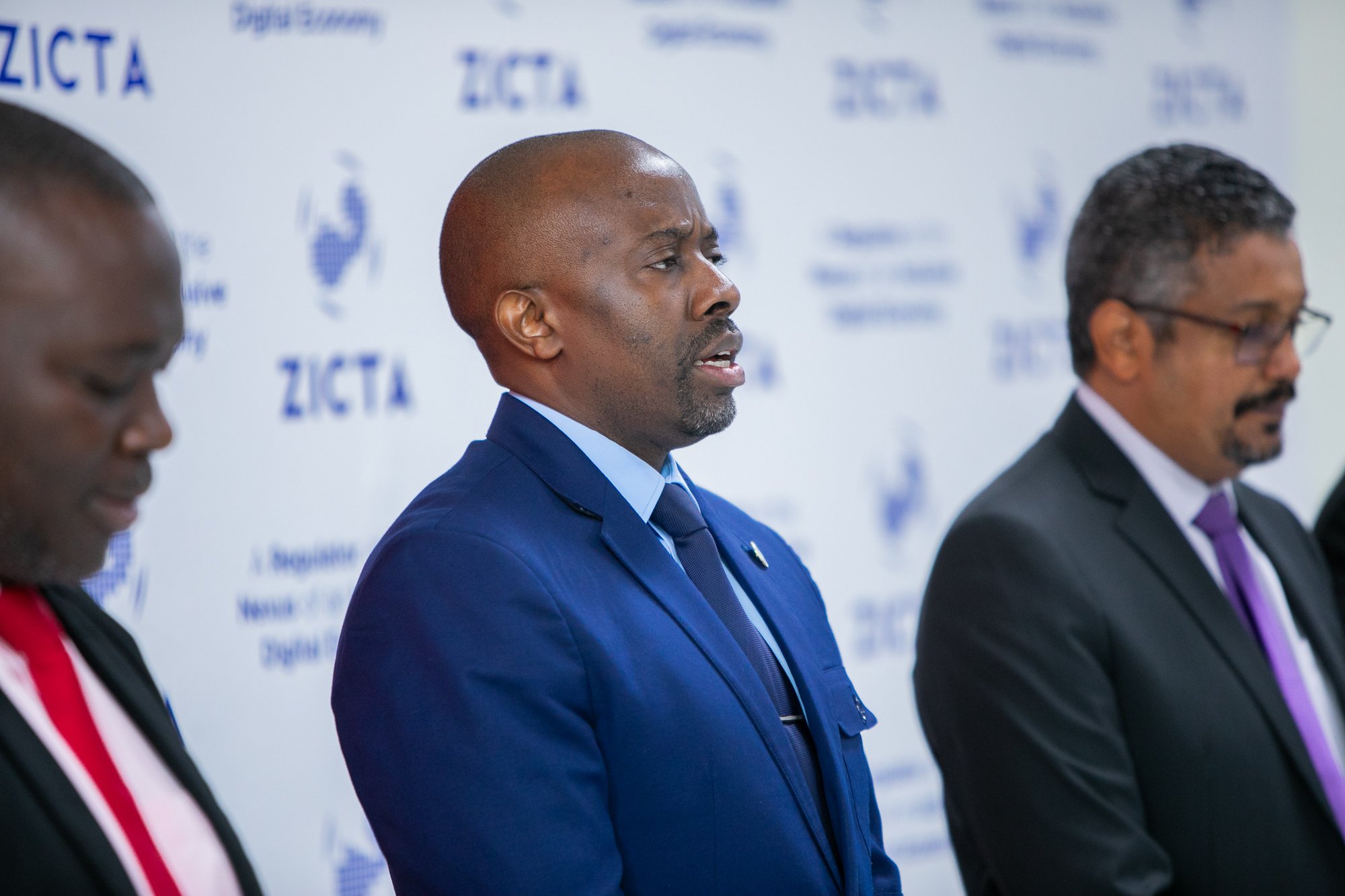 Three men in suits are standing at a table during a formal event or conference, with a backdrop displaying the logo and text of an organization. The focus is on the man in the center who is wearing a blue suit and tie, speaking or listening attentive