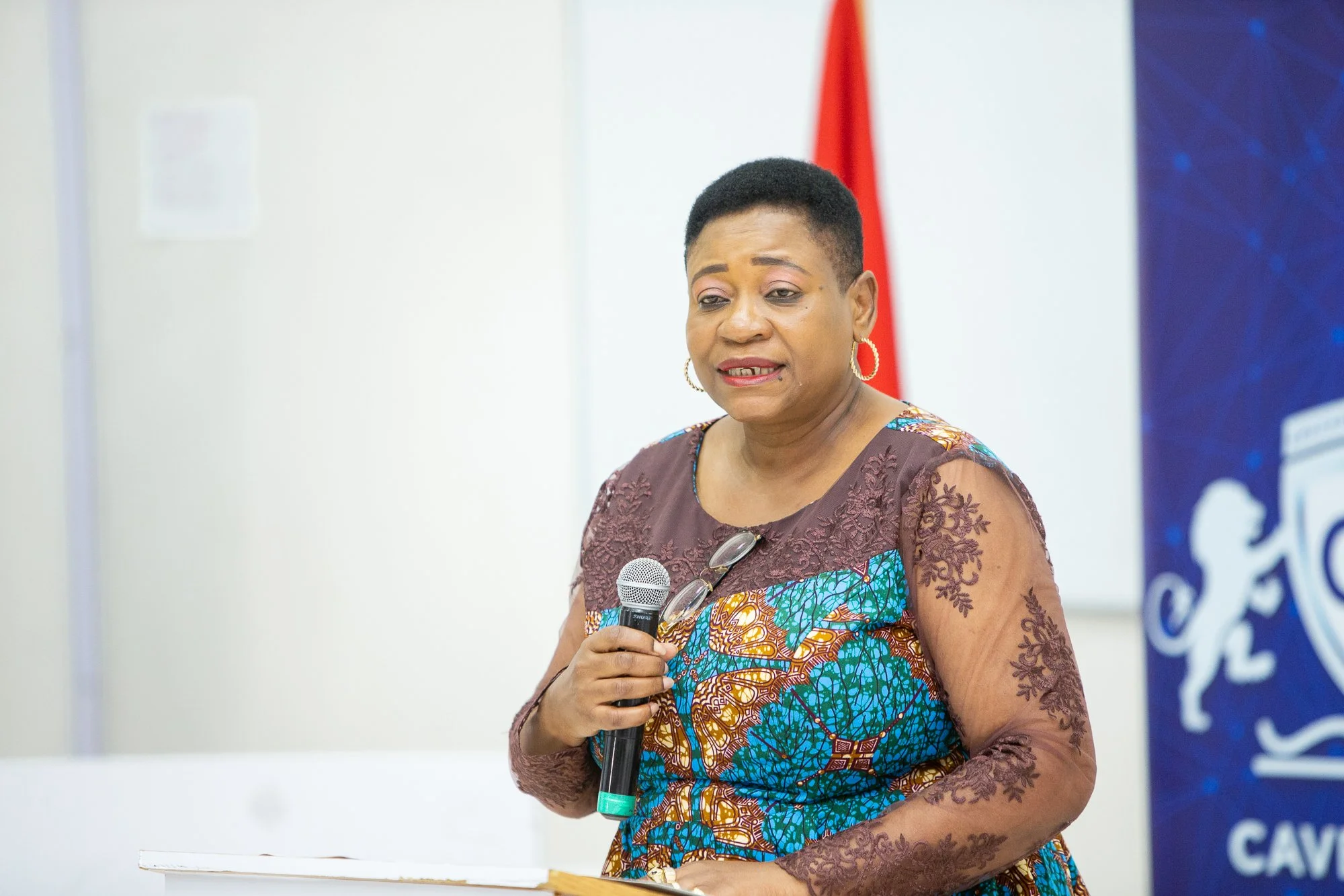 A woman holding a microphone and speaking at an event, wearing a colorful patterned dress with a lace overlay on the sleeves, standing indoors with a white wall and a red flag behind her.