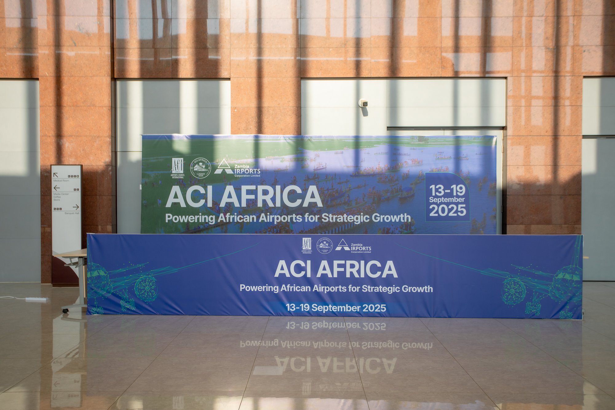 Sign in an airport lobby promoting the ACI Africa conference, scheduled for September 13-19, 2025, with a backdrop of a modern building with large windows and shadows from metal bars.