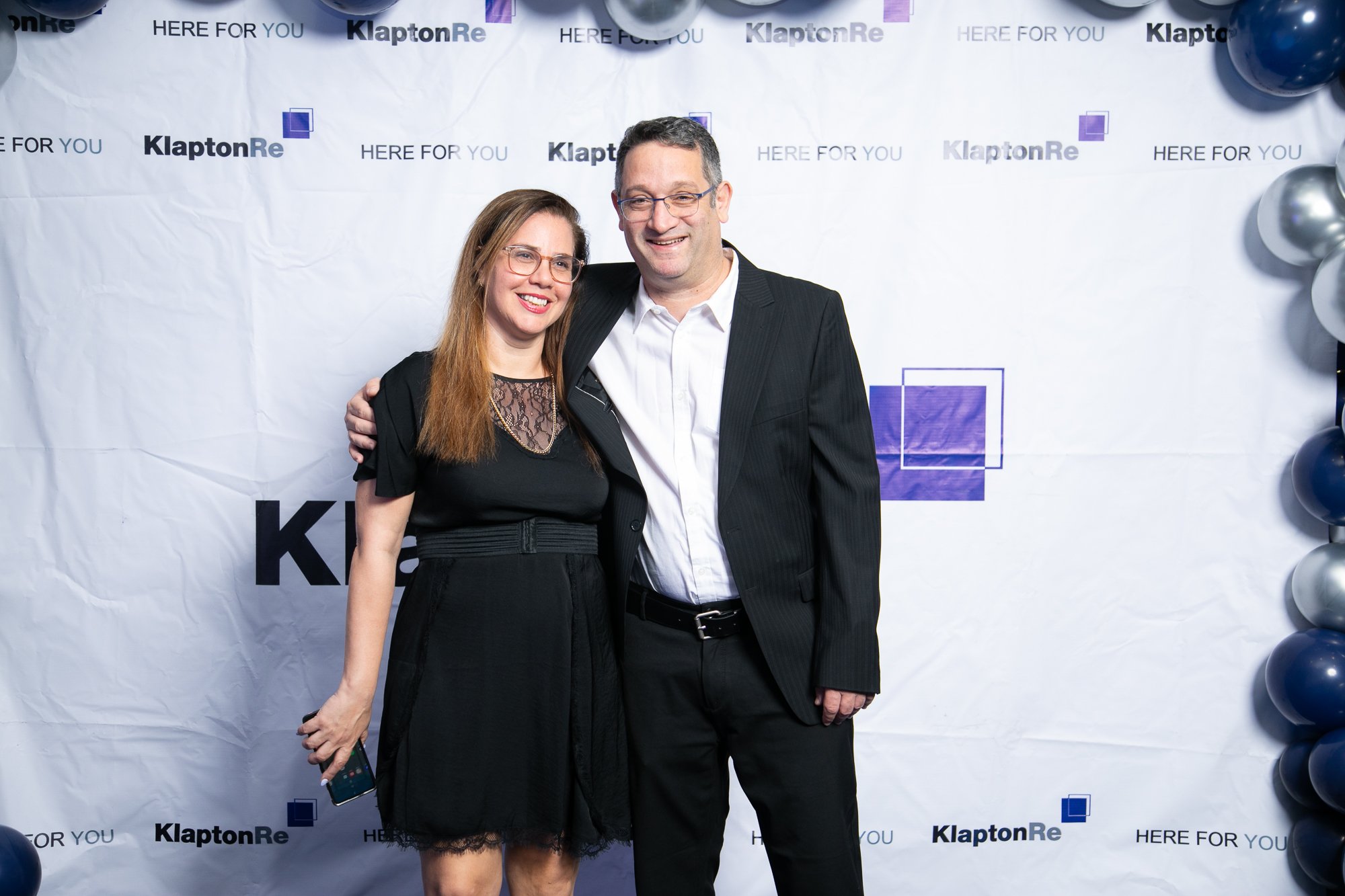 A man and woman standing together at an event with a backdrop featuring the words 'KlaptonRe' and 'here for you,' surrounded by black, blue, and white balloons.