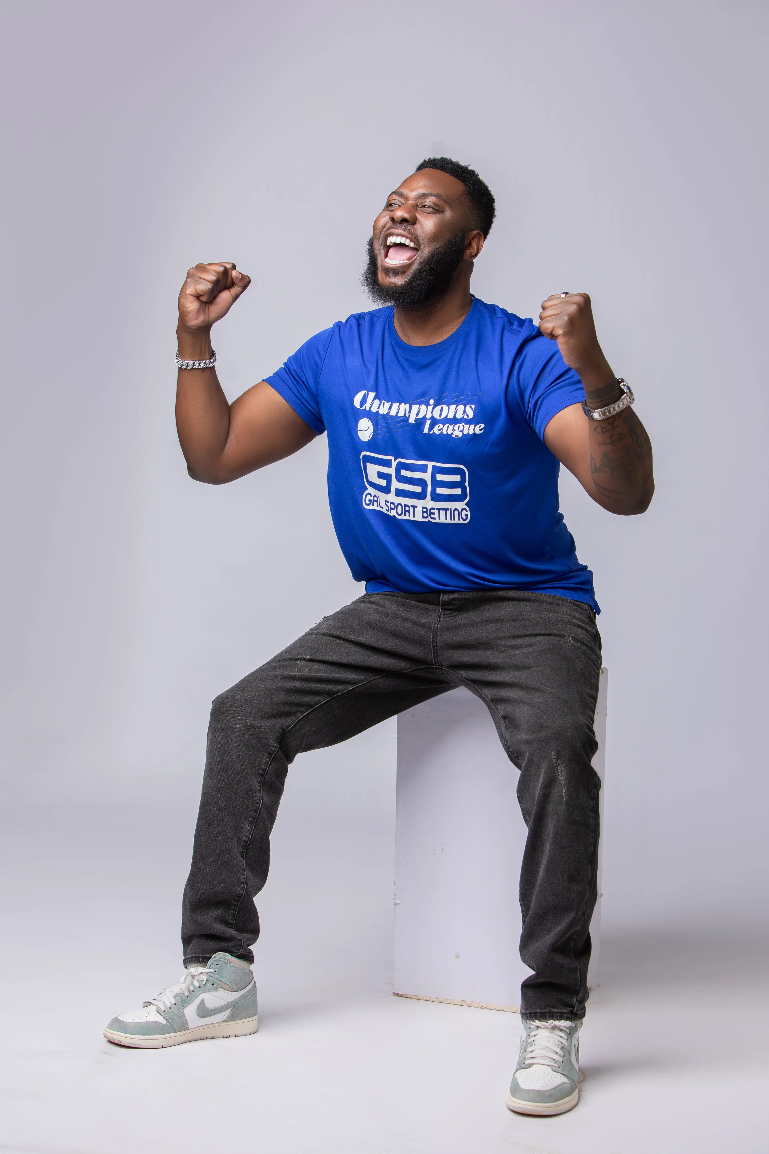 Slap Dee with a beard celebrating with fists raised, wearing a blue 'Champions League' and 'GSP GAL SPORT BETTING' t-shirt, gray jeans, and white sneakers, against a plain white background.