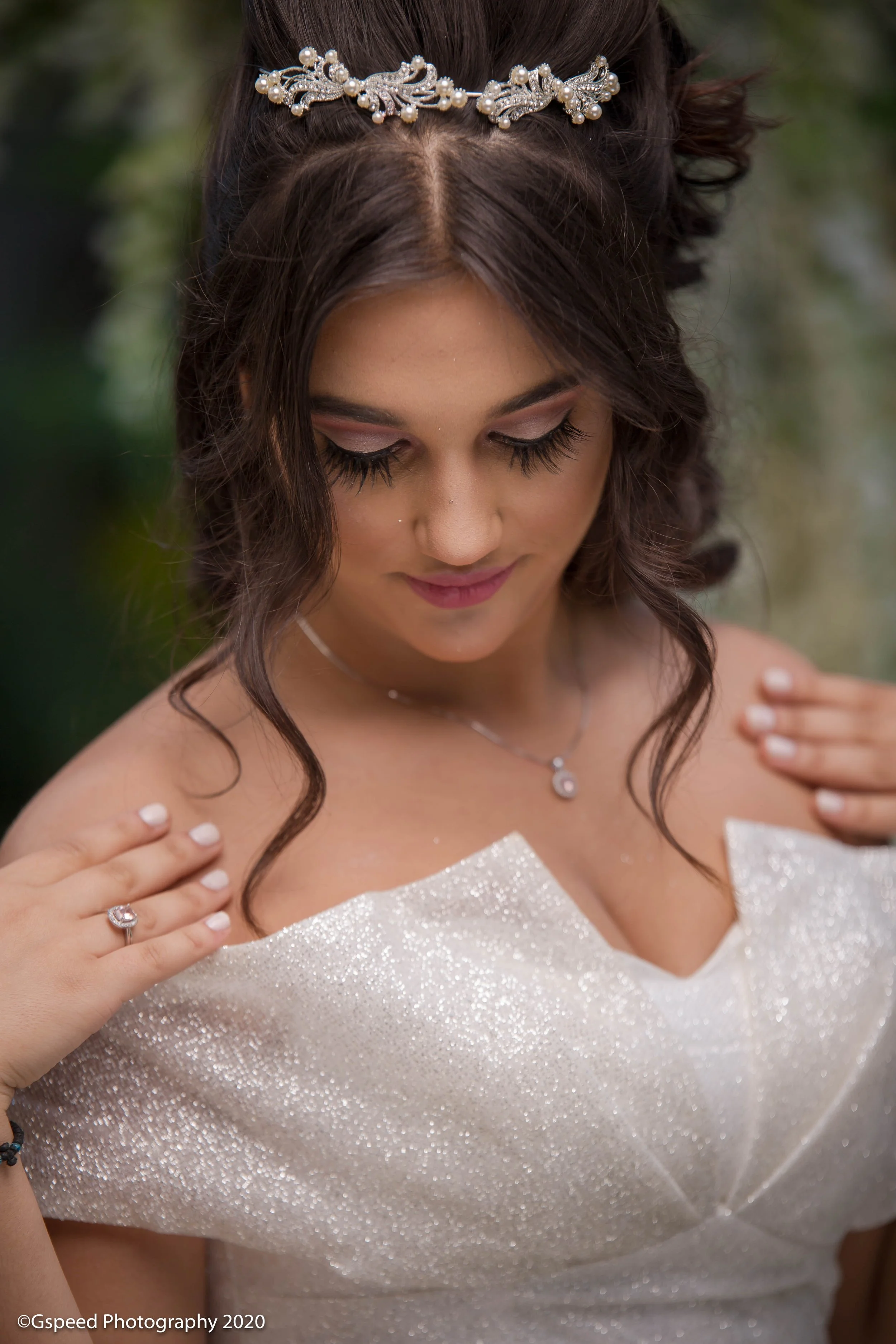 A close-up of a bride wearing a pearl and crystal tiara, with dark wavy hair, and a sparkly off-shoulder wedding dress. She has makeup with soft pink lipstick and long lashes, and is gently touching her shoulder with both hands, adorned with a ring a