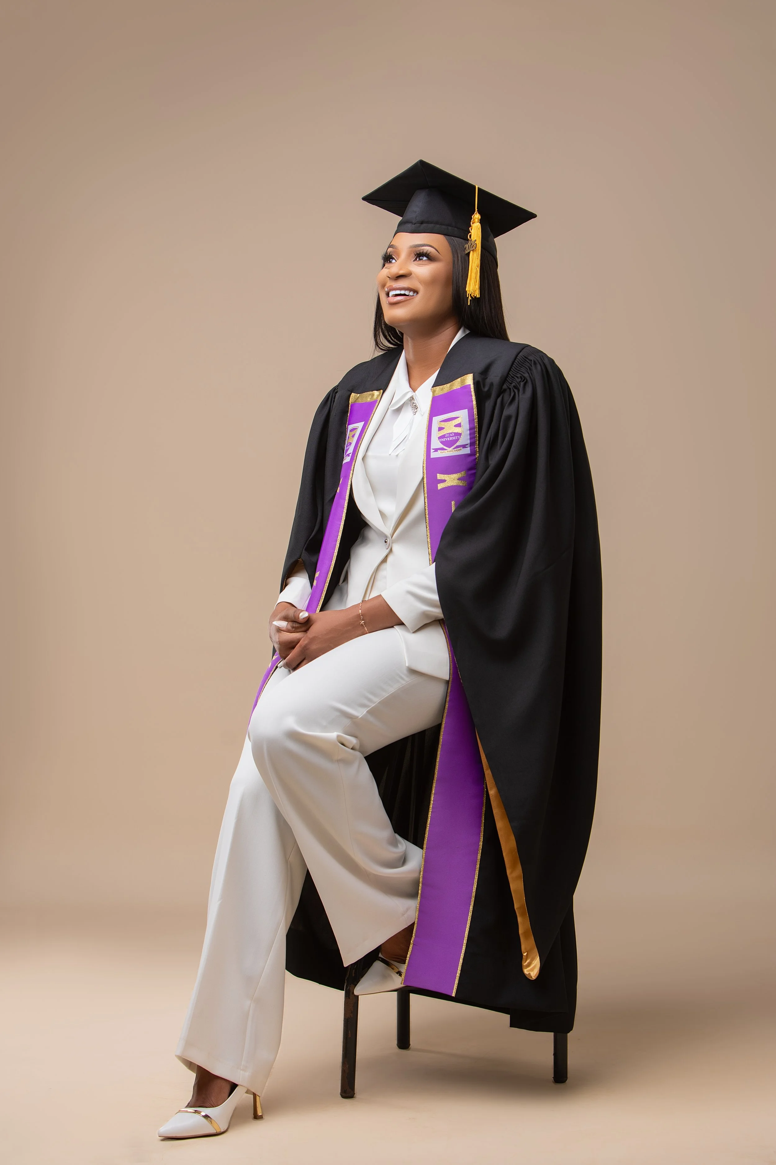 A woman dressed in a white suit and white high heels, wearing a black graduation cap and gown with purple and gold accents, sitting on a stool with one leg crossed over the other, smiling and looking upward against a plain beige background.