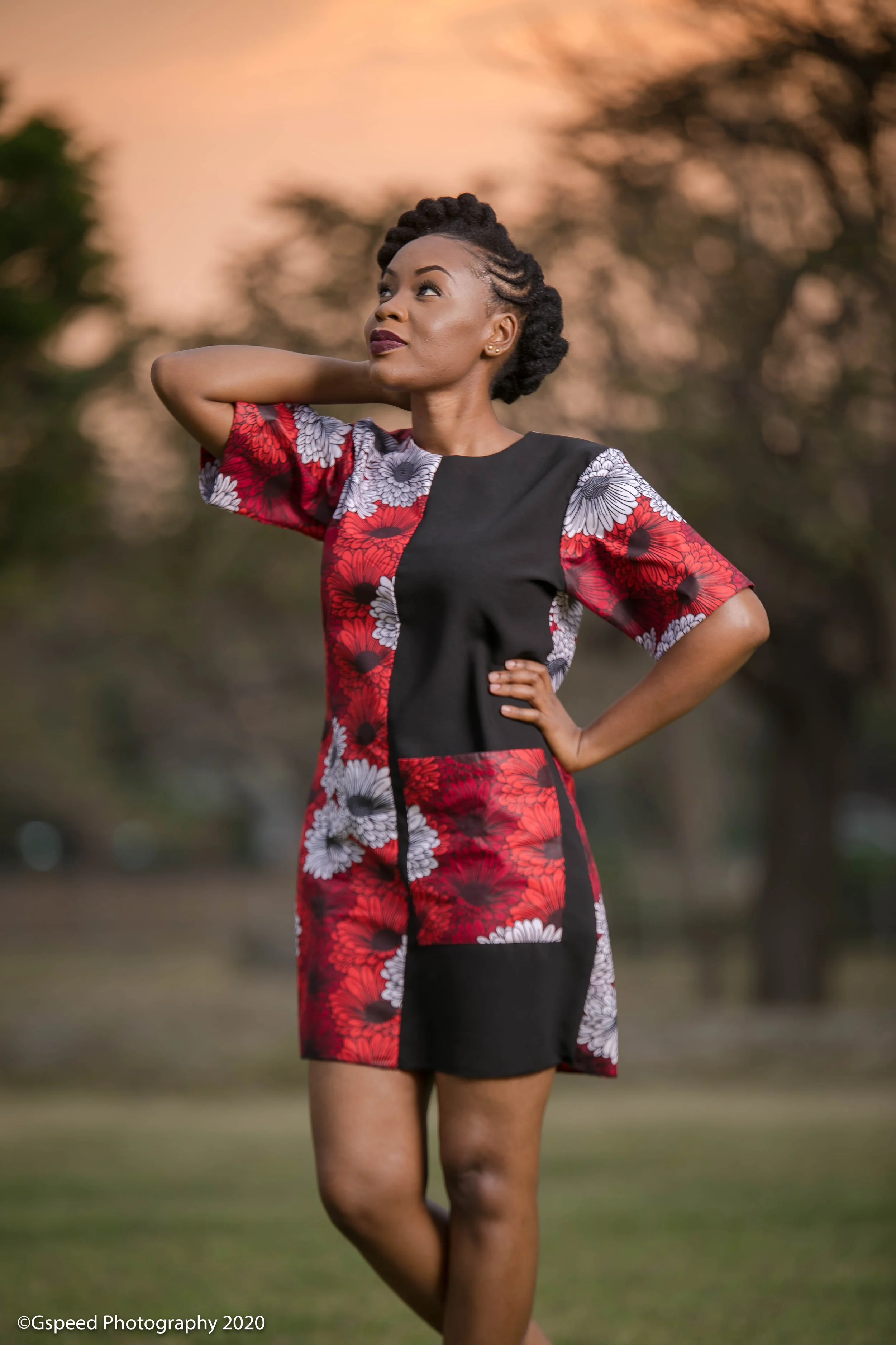 A woman with dark hair styled in twists, standing outdoors during sunset, wearing a dress with a floral pattern in red, black, and white, with one hand on her hip and the other behind her head, looking to the side.