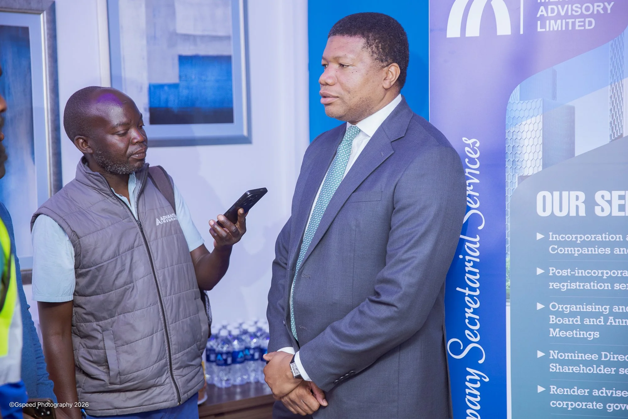 Three men engaged in conversation at a professional event. One man in a suit, another in a gray vest, and a third partially visible on the left. A corporate banner with blue and purple colors and text is in the background.