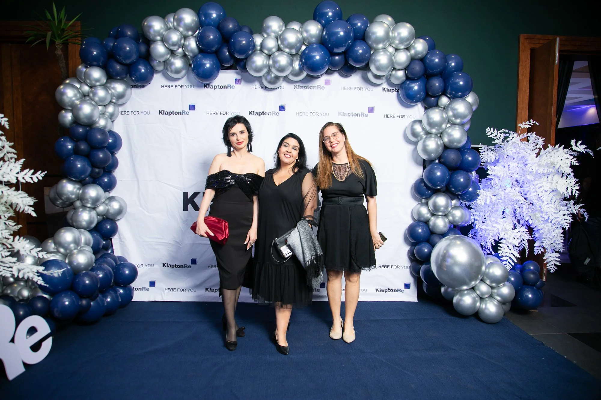 Three women dressed in formal black attire standing in front of a white backdrop with 'KlaptonRe' and 'Here for You' logos, framed by arching blue and silver balloons and white decorative plants, at what appears to be a celebratory or corporate event