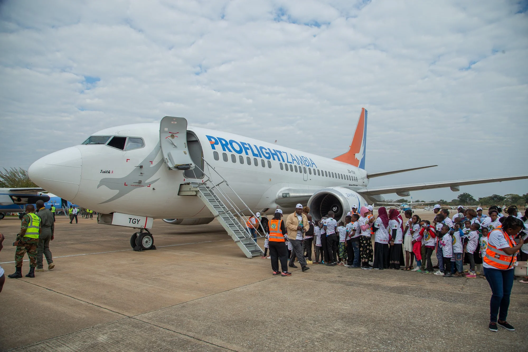 A large airplane with the words 'Proflight Zambia' on its side is parked on the tarmac during an event. A staircase is leading into the plane's open door. Many children and adults are gathered around, some in safety vests, with a cloudy sky overhead.