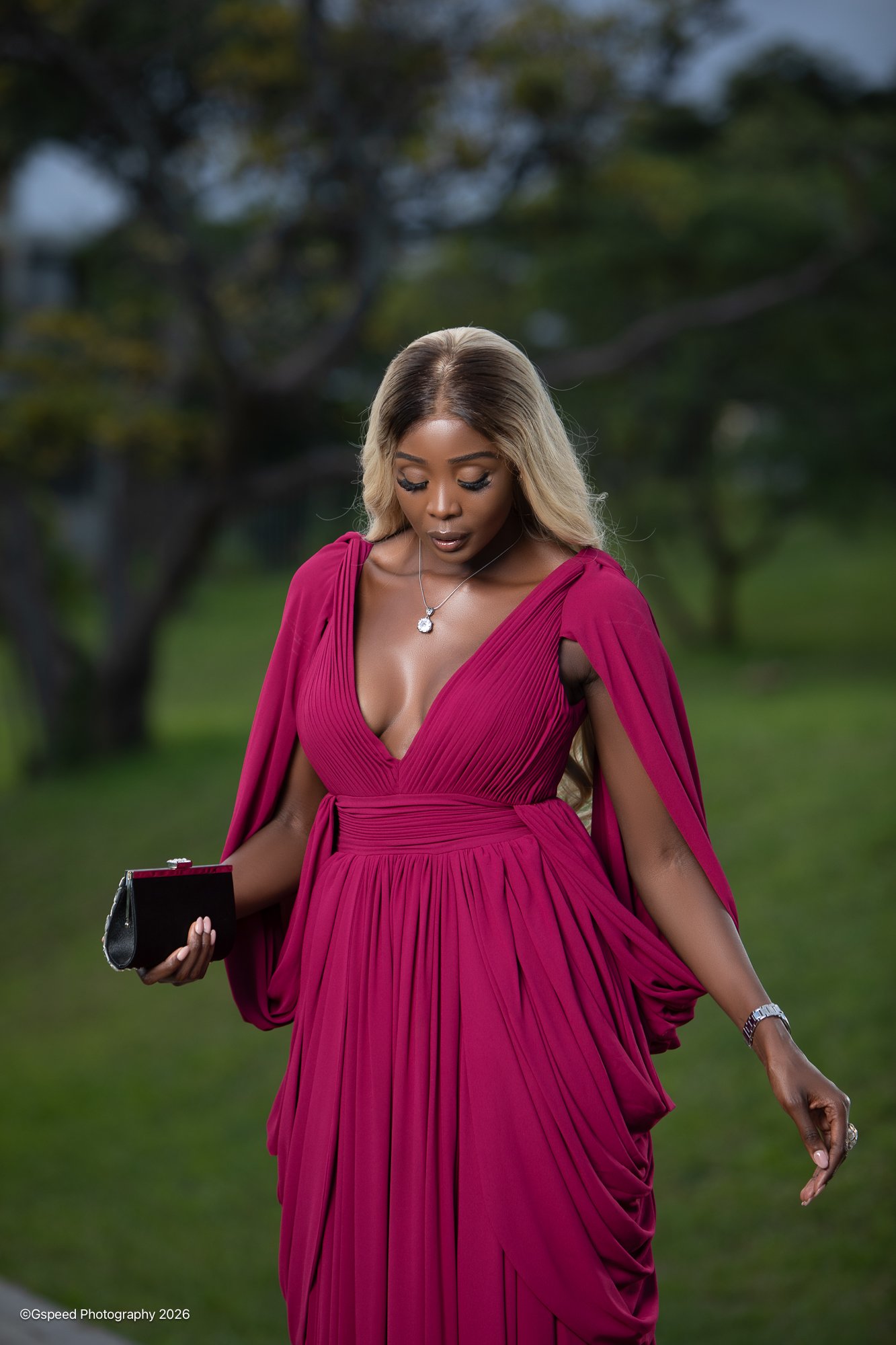 A woman in a flowing pink gown outdoors, looking down and holding a small black clutch purse, with a blurred background of trees and grass.