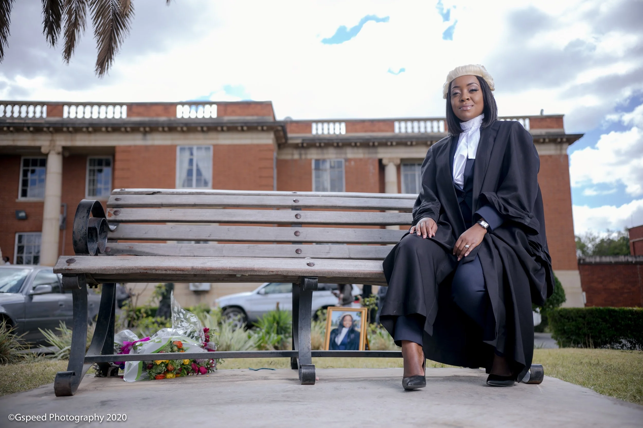 A woman dressed in a black graduation gown and cap sitting on a park bench during a memorial or funeral service. Flowers and a framed photo are placed on the ground nearby, and a building and parked cars are visible in the background under a cloudy s