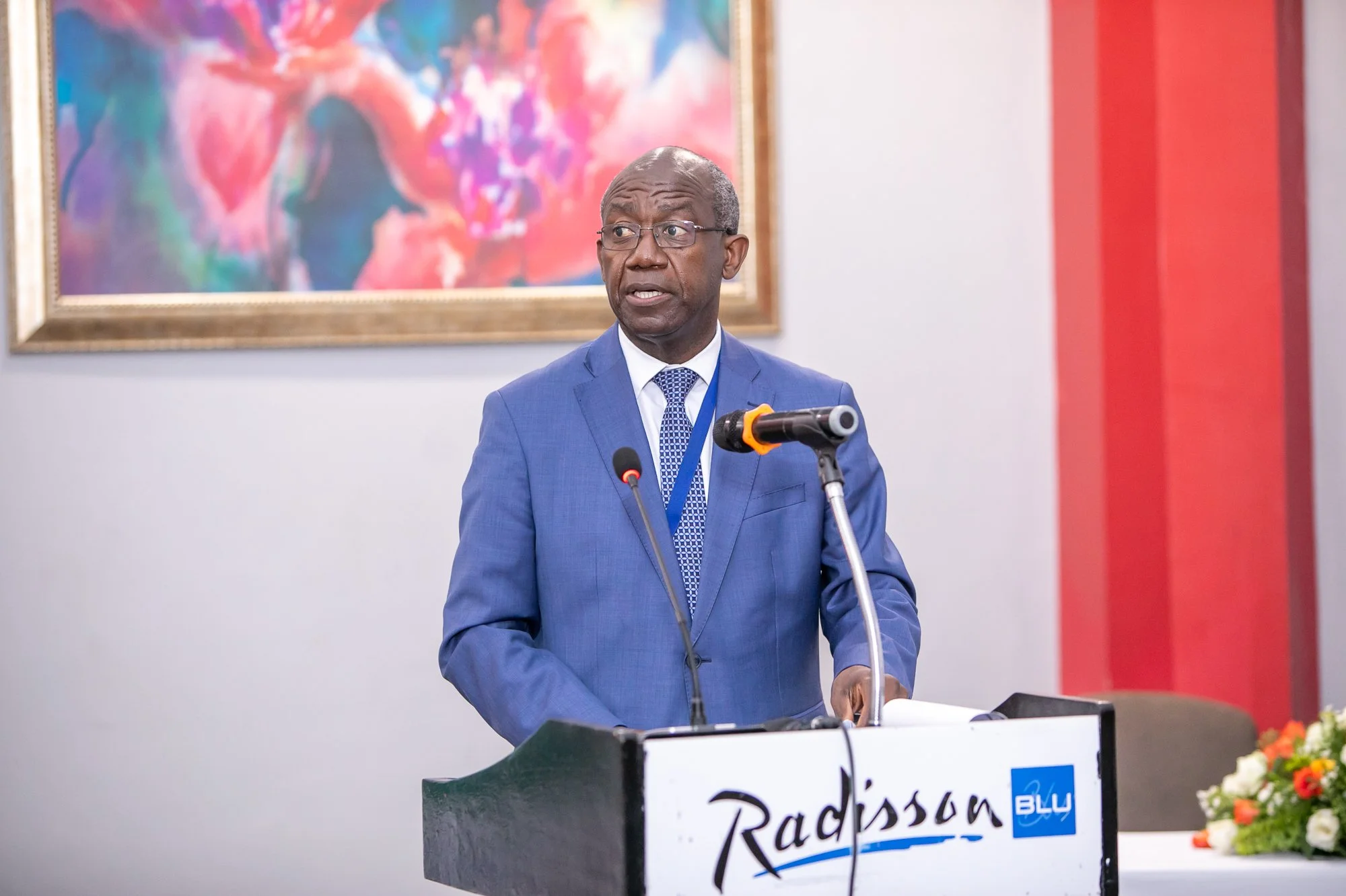 A man in a blue suit and glasses is standing at a podium giving a speech at a conference. There is a large colorful painting on the wall behind him.