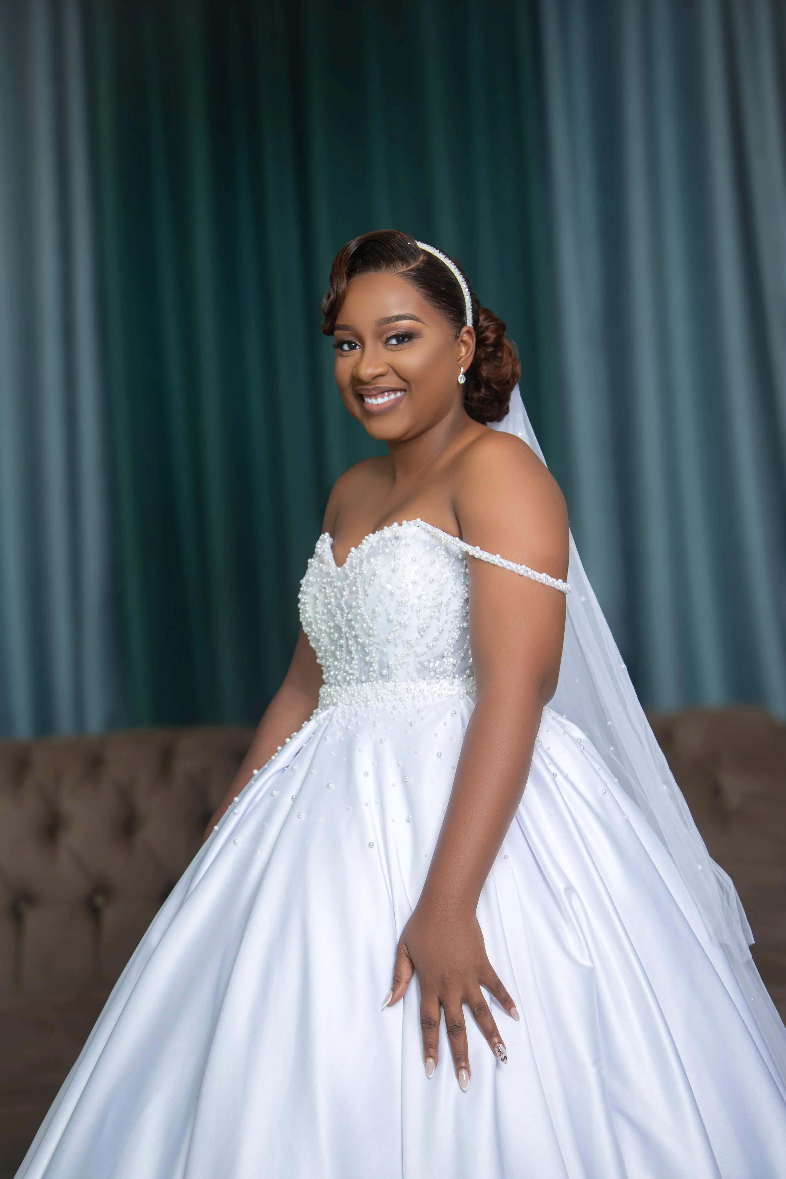 A smiling bride in a white wedding gown with beaded details against a green curtain background.