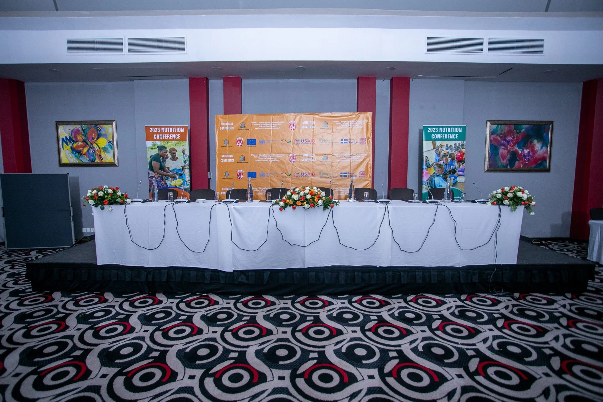 A conference room with a long table covered with a white tablecloth and decorated with floral arrangements. Behind the table, there is a large orange banner with logos and text about the 2023 Nutrition Conference, along with two vertical banners on e