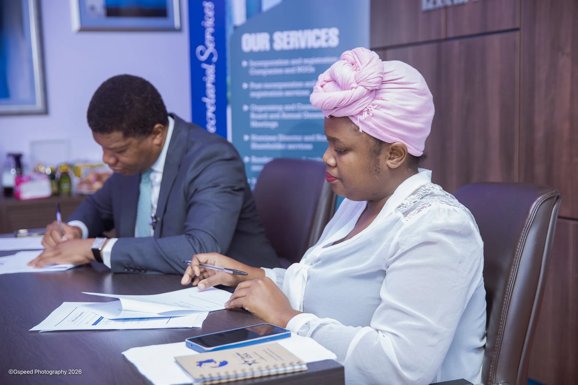 Two individuals sitting at a conference table during a meeting, both writing on papers. The person on the left is a man in a suit, and the person on the right is a woman with a pink headwrap and white top. The background features a blue sign with 'Ou