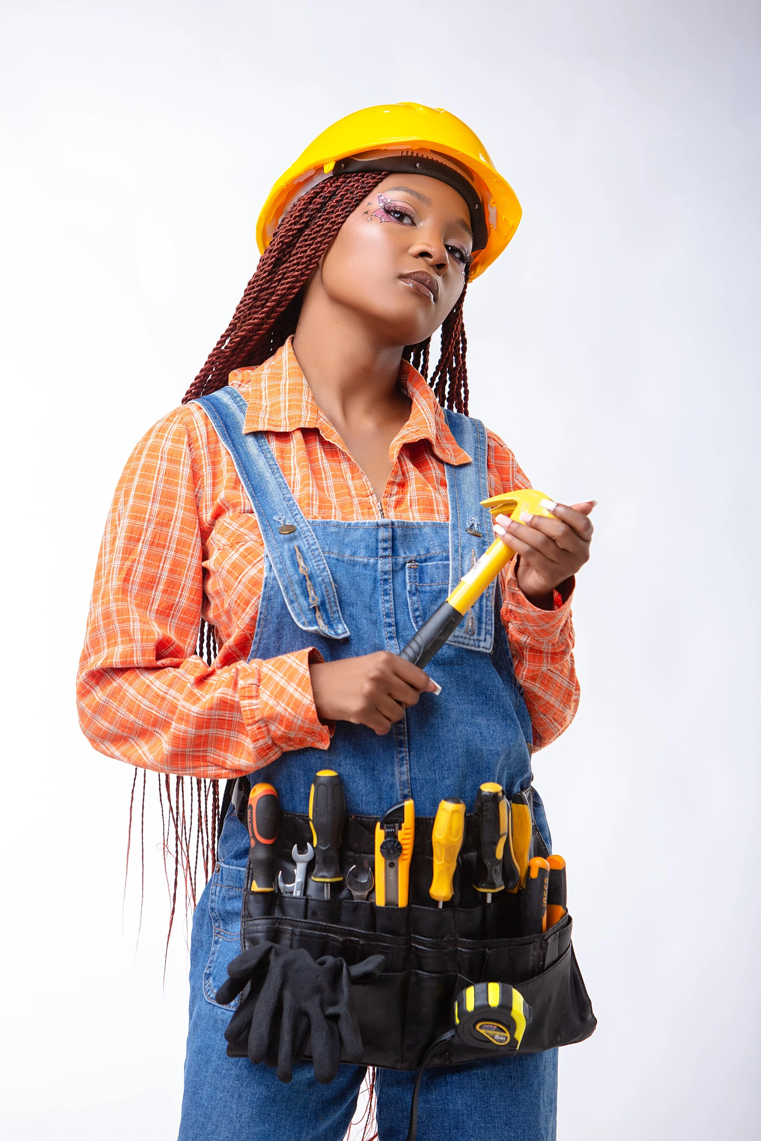 A woman wearing a yellow construction helmet, orange plaid shirt, and denim overalls, holding a screwdriver, with various tools in a tool belt around her waist.