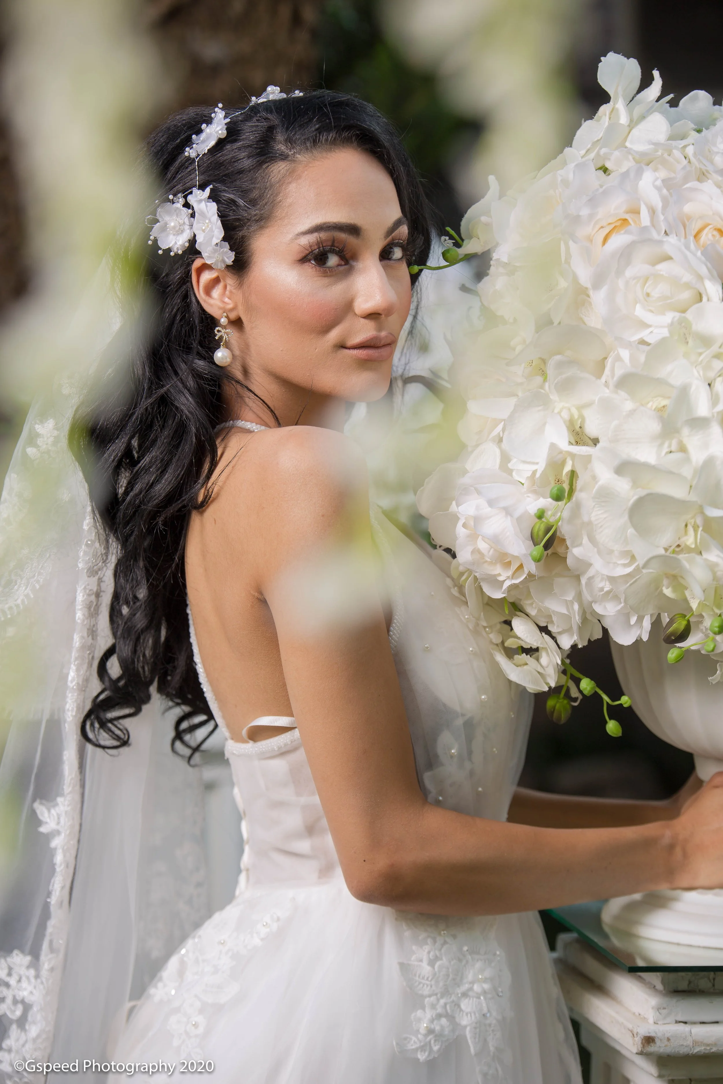 A woman in a white wedding dress holding a large bouquet of white flowers, with decorated hair and earrings, in a setting that appears to be a florist or bridal shop.