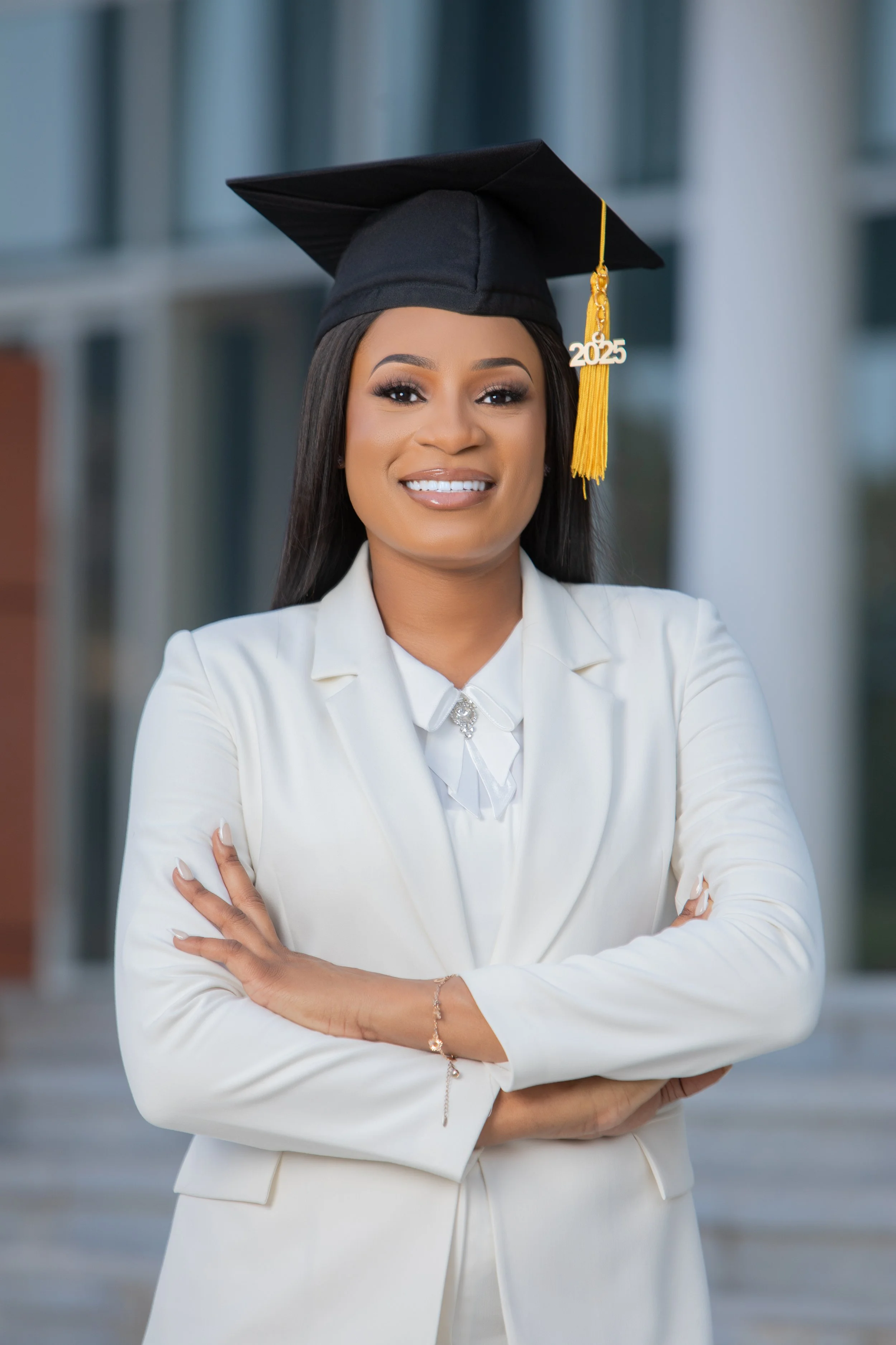 A young woman wearing a white blazer and a white blouse with a small silver decorative pin, smiling with arms crossed, standing outdoors in front of a modern building with glass and concrete columns. She is wearing a black graduation cap with a yello