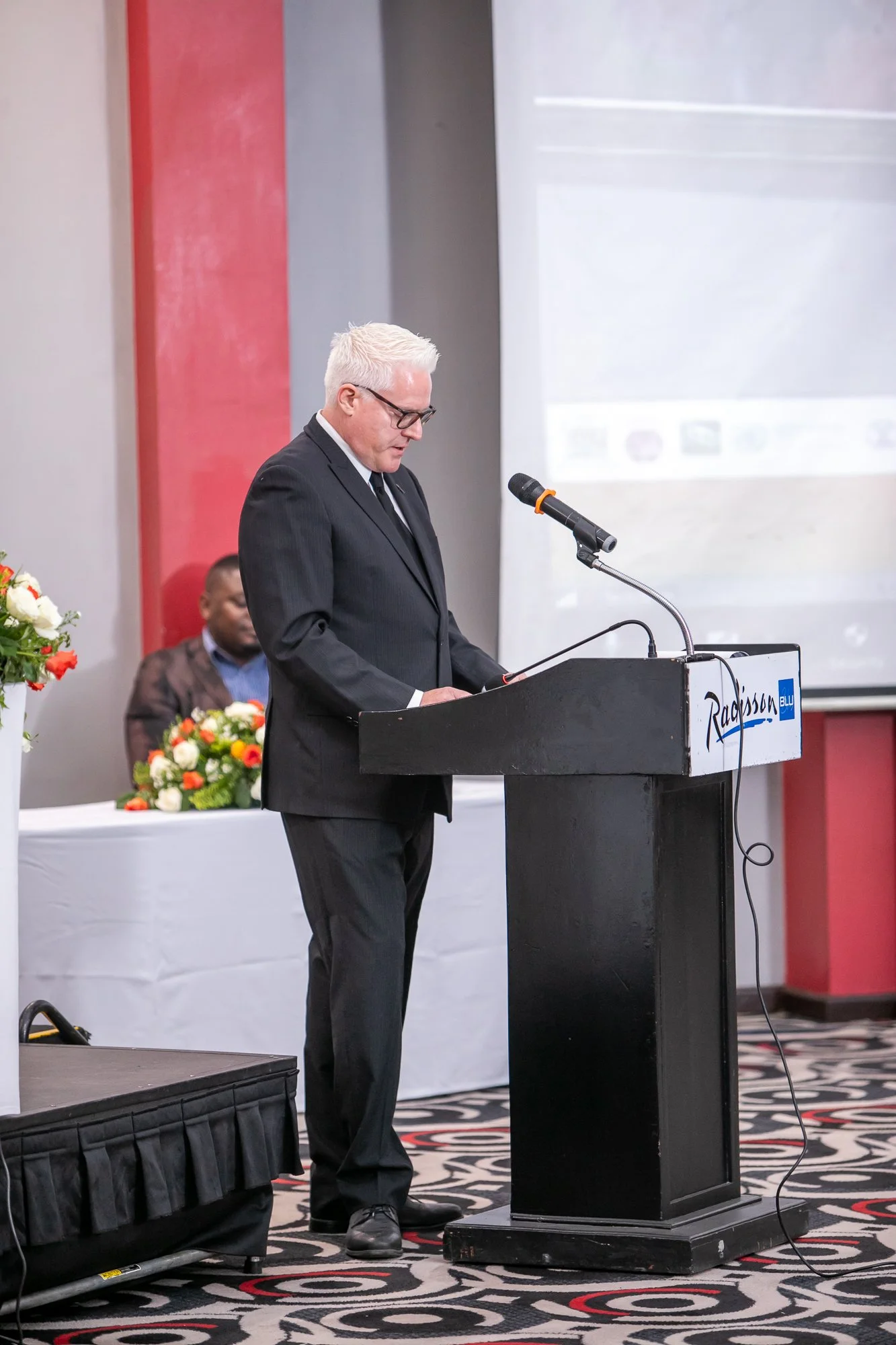 A man with white hair and glasses in a black suit stands at a podium with a microphone, reading or speaking during a formal event at a conference room decorated with flowers and a patterned carpet.