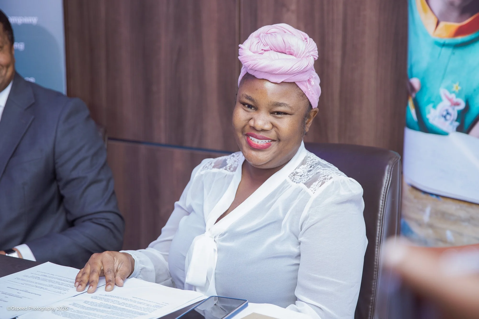 A woman with a pink headwrap, smiling, seated at a table with papers in front of her, wearing a white blouse with lace details on the shoulders.