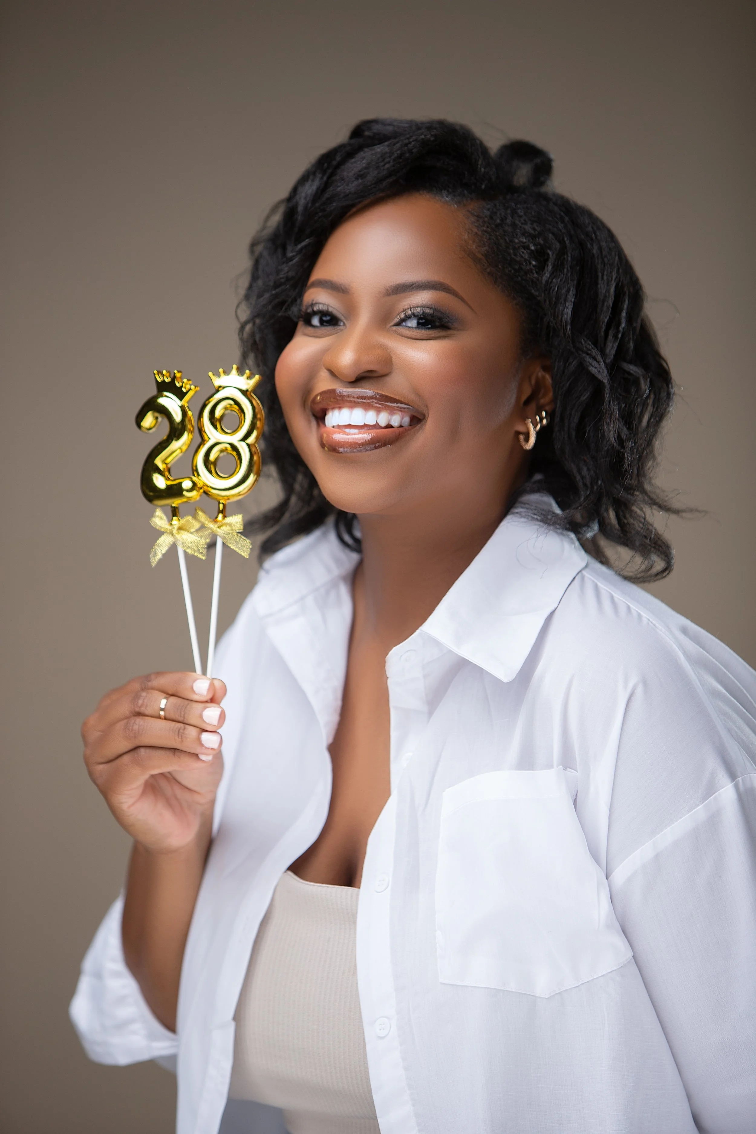 birthday studio photoshoot of woman with dark curly hair smiling and holding gold balloons that spell out 28, wearing a white shirt and beige tank top against a neutral background.