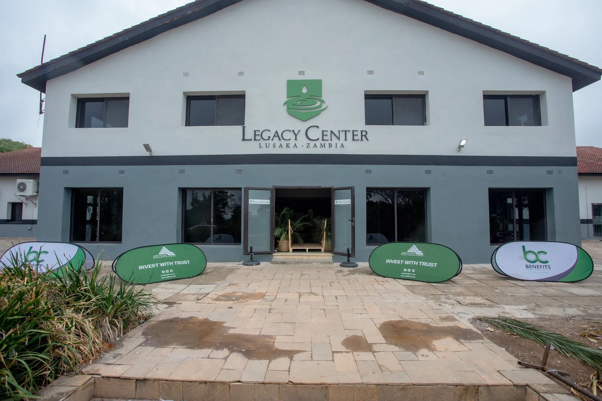 Front view of a two-story building labeled "Legacy Center Lusaka, Zambia" with a green water drop logo. Four green and white promotional banners are placed in front, reading "Invest with Trust" and other messages, with some plants and a paved pathway