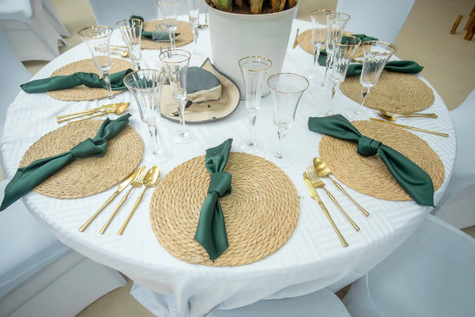 Round dining table set with woven straw placemats, green cloth napkins tied at the top, gold flatware, and empty wine glasses with gold rims, with a large white planter in the center.