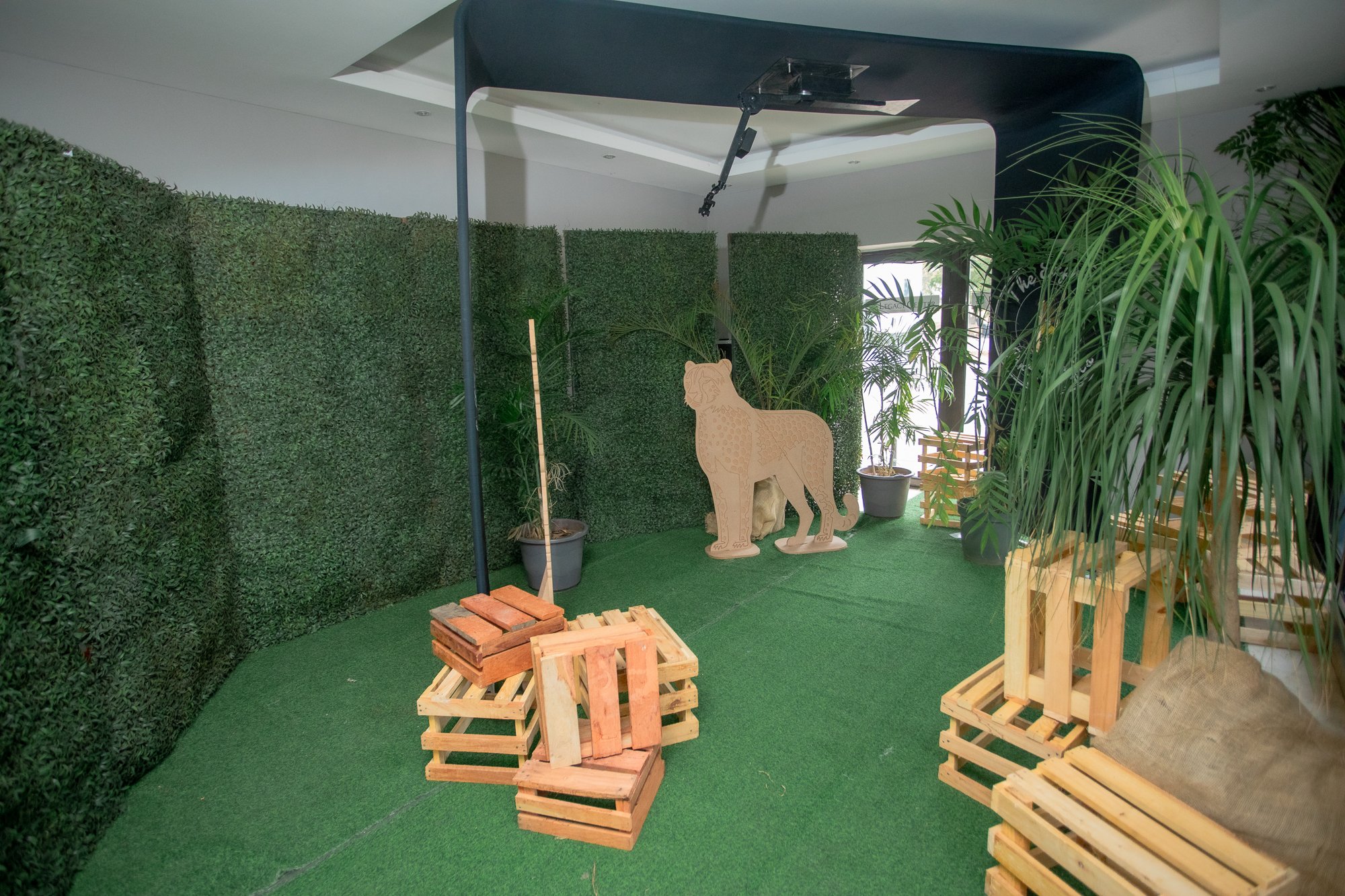 Indoor space decorated with green artificial turf and lush potted plants. Wooden pallets are stacked and arranged as furniture. A cardboard cut-out of a lion stands in front of a hedge. Overhead, there is a black structure with mechanical parts hangi
