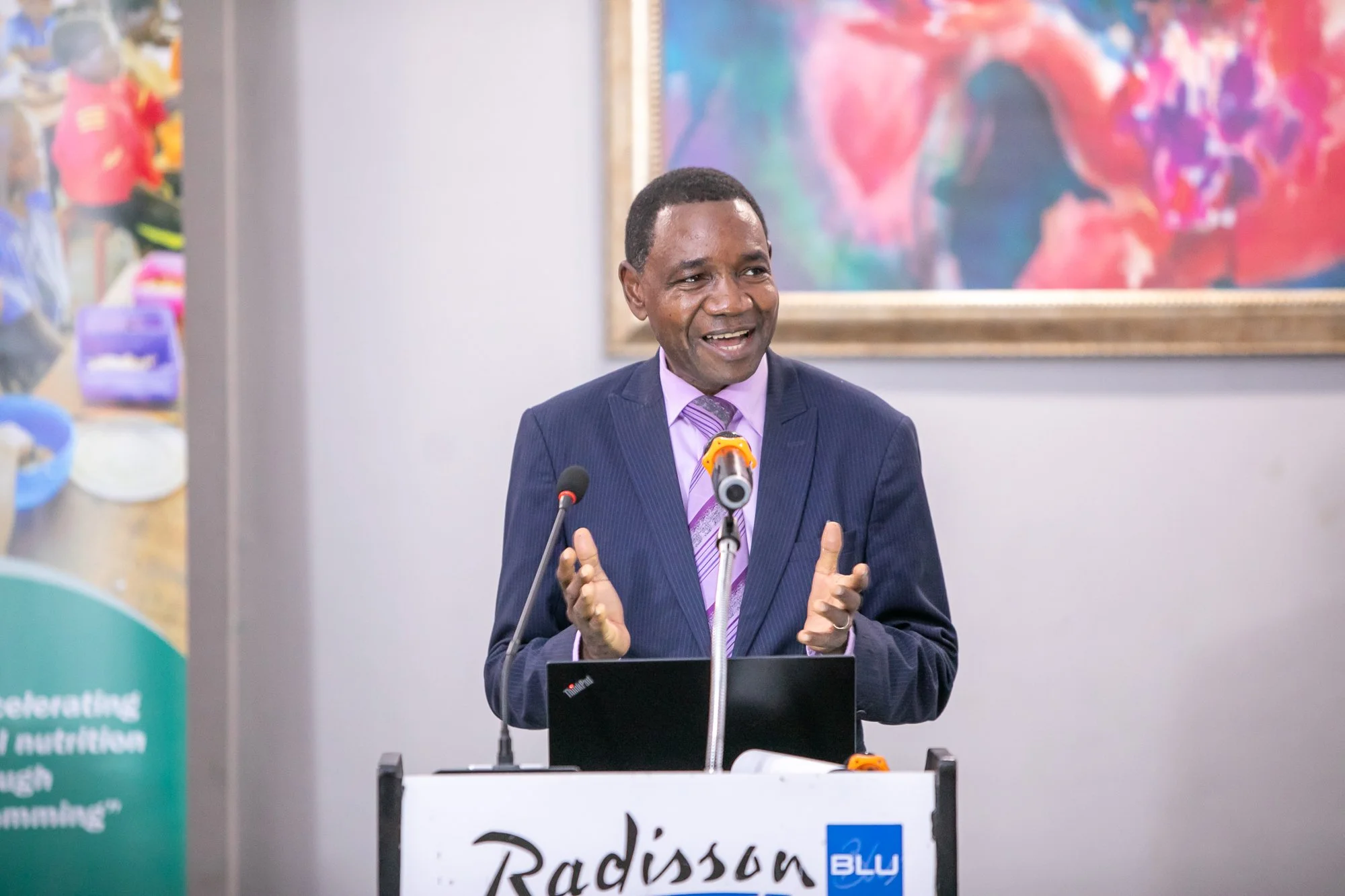 A man in a dark suit and purple shirt speaking at a podium with a sign that reads 'Radisson Blu.' Behind him is colorful abstract artwork on the wall.