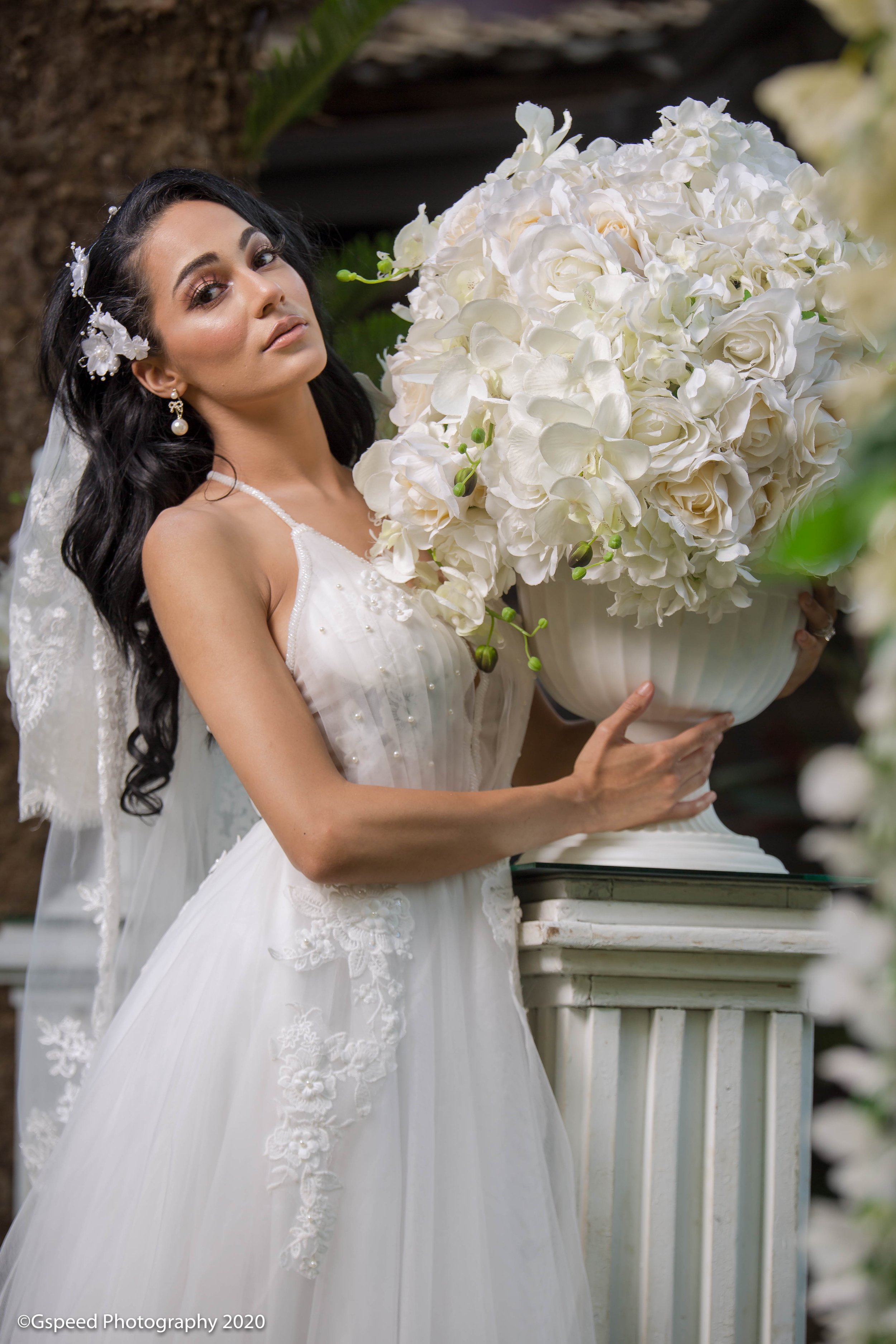 A bride with long black wavy hair, wearing pearl earrings and a white dress with lace and embroidery, holding a large bouquet of white flowers, standing by a white wooden railing in an outdoor setting.