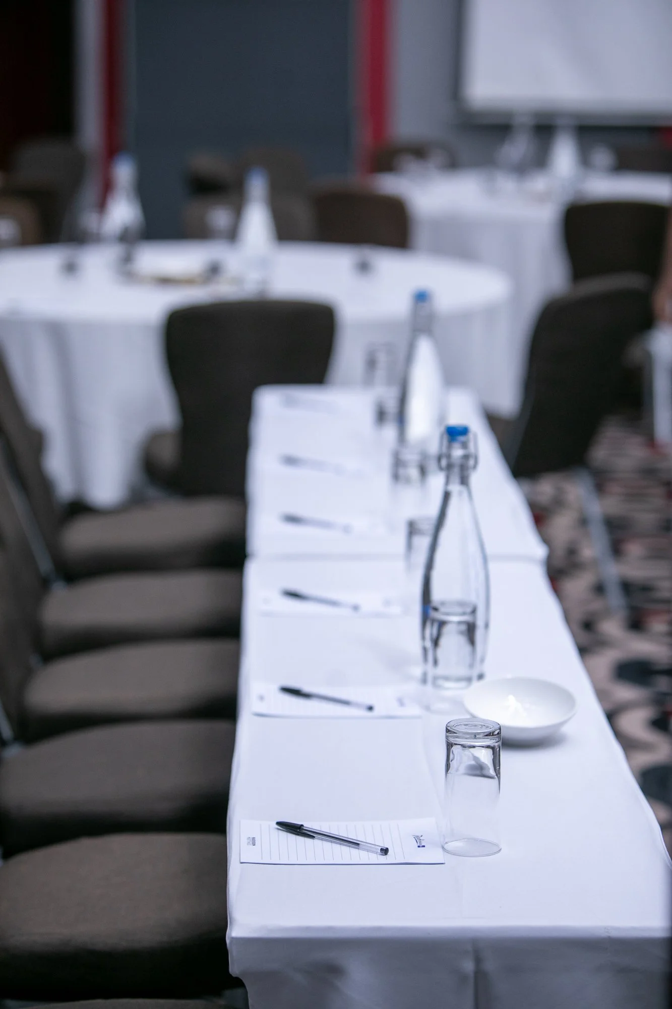 Conference room with round tables covered with white tablecloths, each set with water bottles, glasses, and notepads, and surrounded by black chairs.