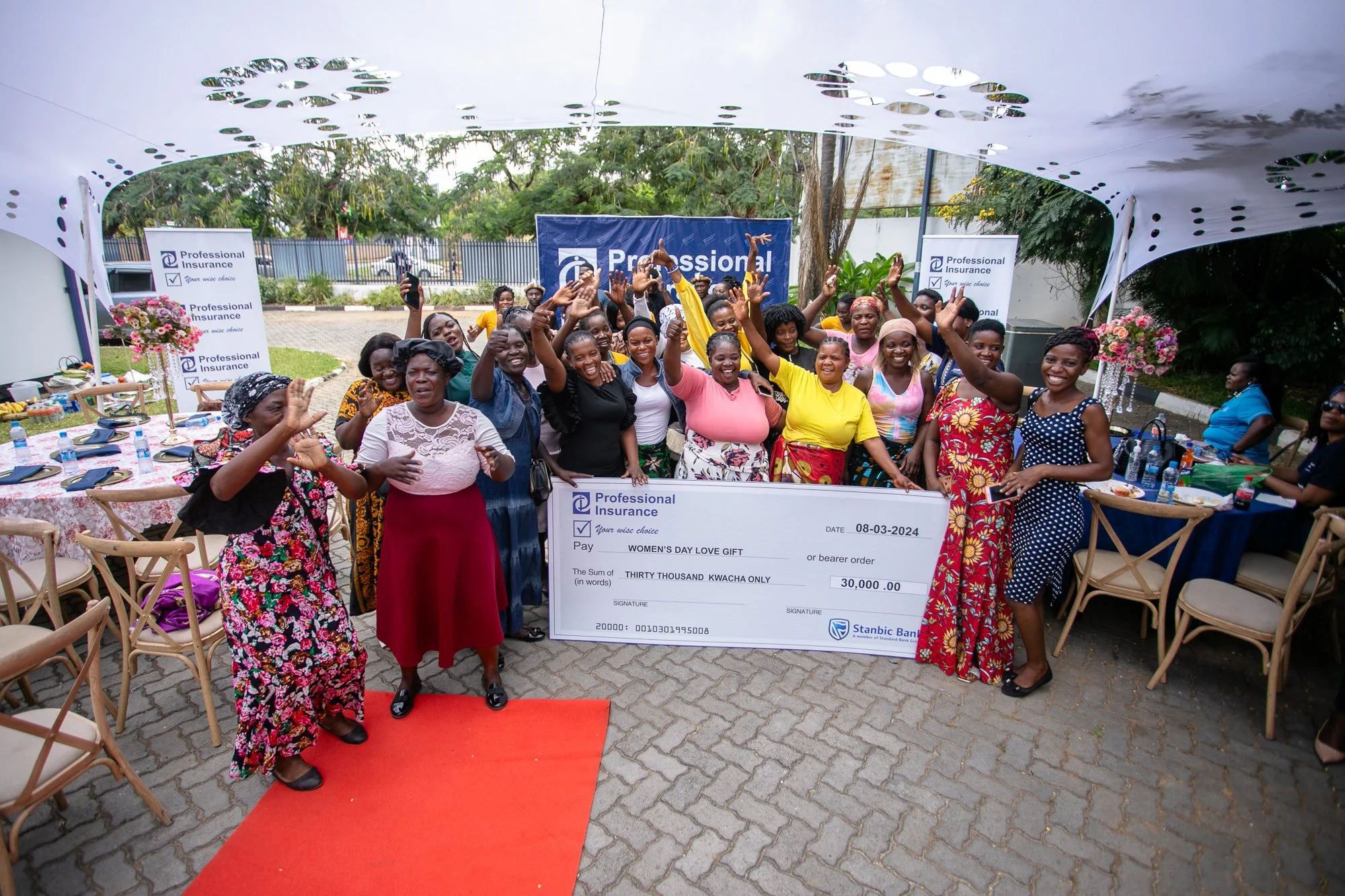 A group of women smiling and waving at a Women’s Day celebration event, standing behind a large ceremonial check for 30,000 Kenyan Shillings from Stanbic Bank, outdoors under a decorative tent.