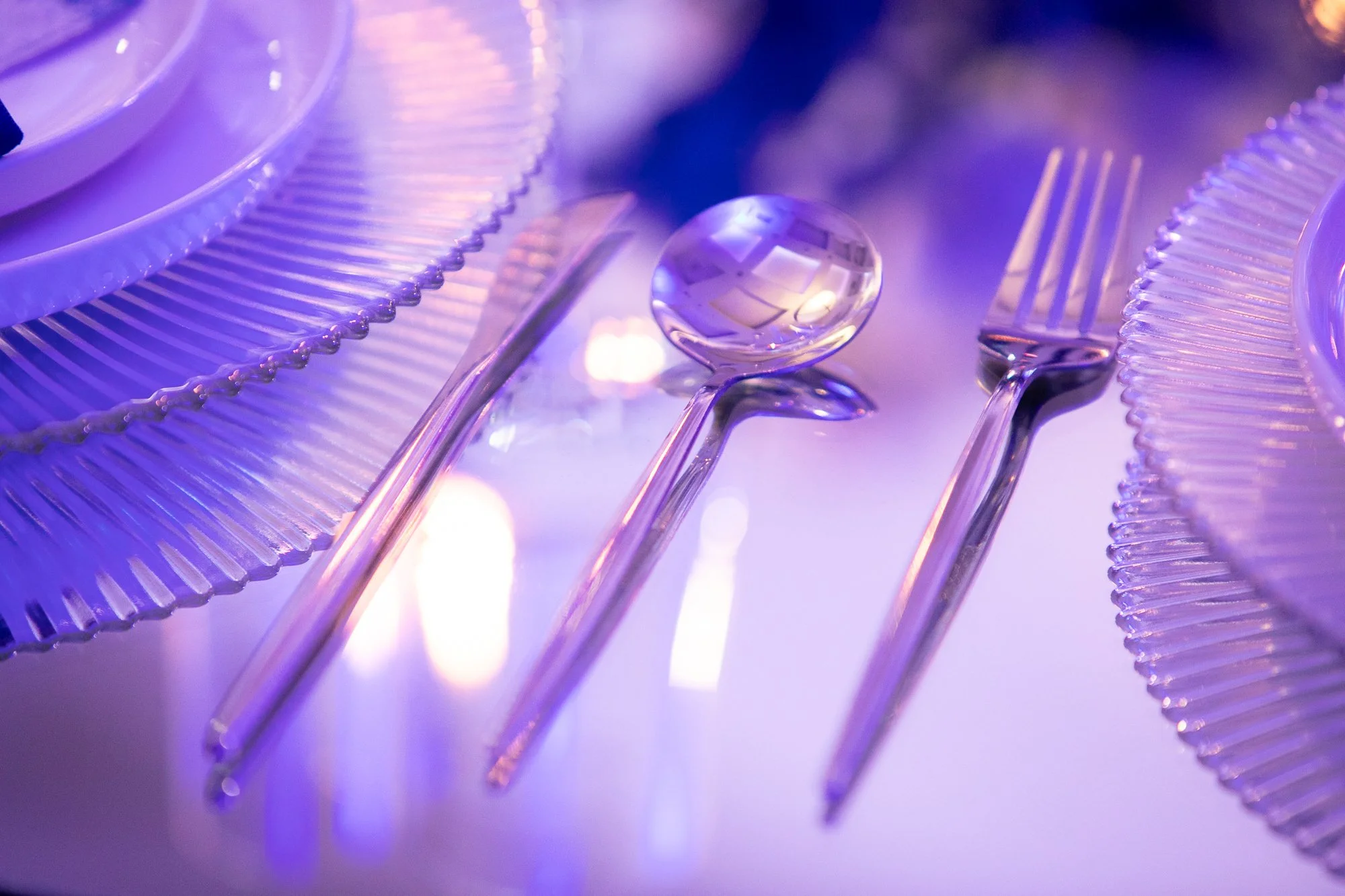 A close-up of transparent plastic cutlery, including a spoon and forks, with decorative plates around them. The scene is illuminated with purple and blue lighting, creating a reflective and vibrant atmosphere.