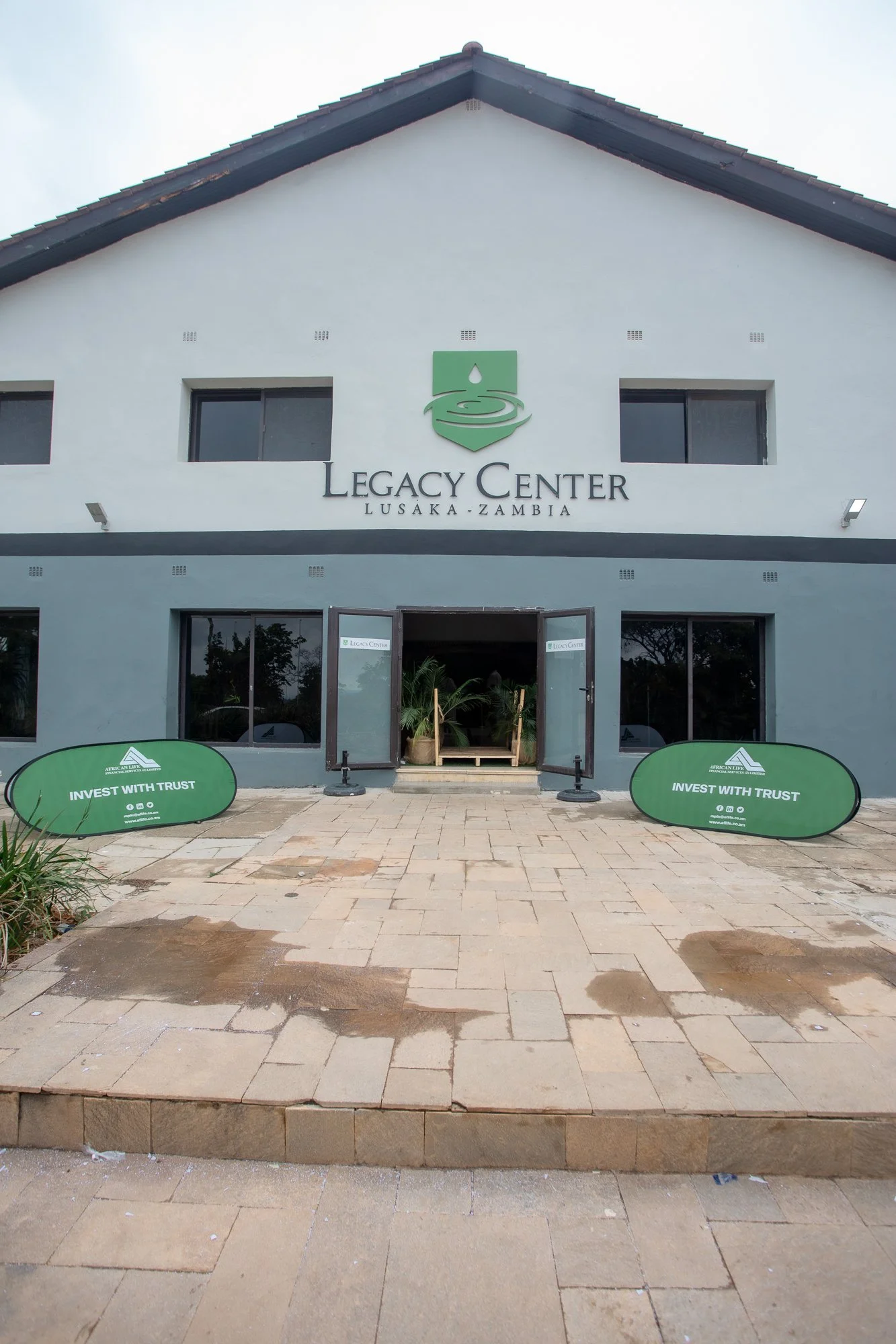 Front view of the Legacy Center in Lusaka, Zambia, with the main entrance, two windows above the door, green banners on either side of the pathway that read 'Invest with Trust,' and a logo showing a water drop above a hand.